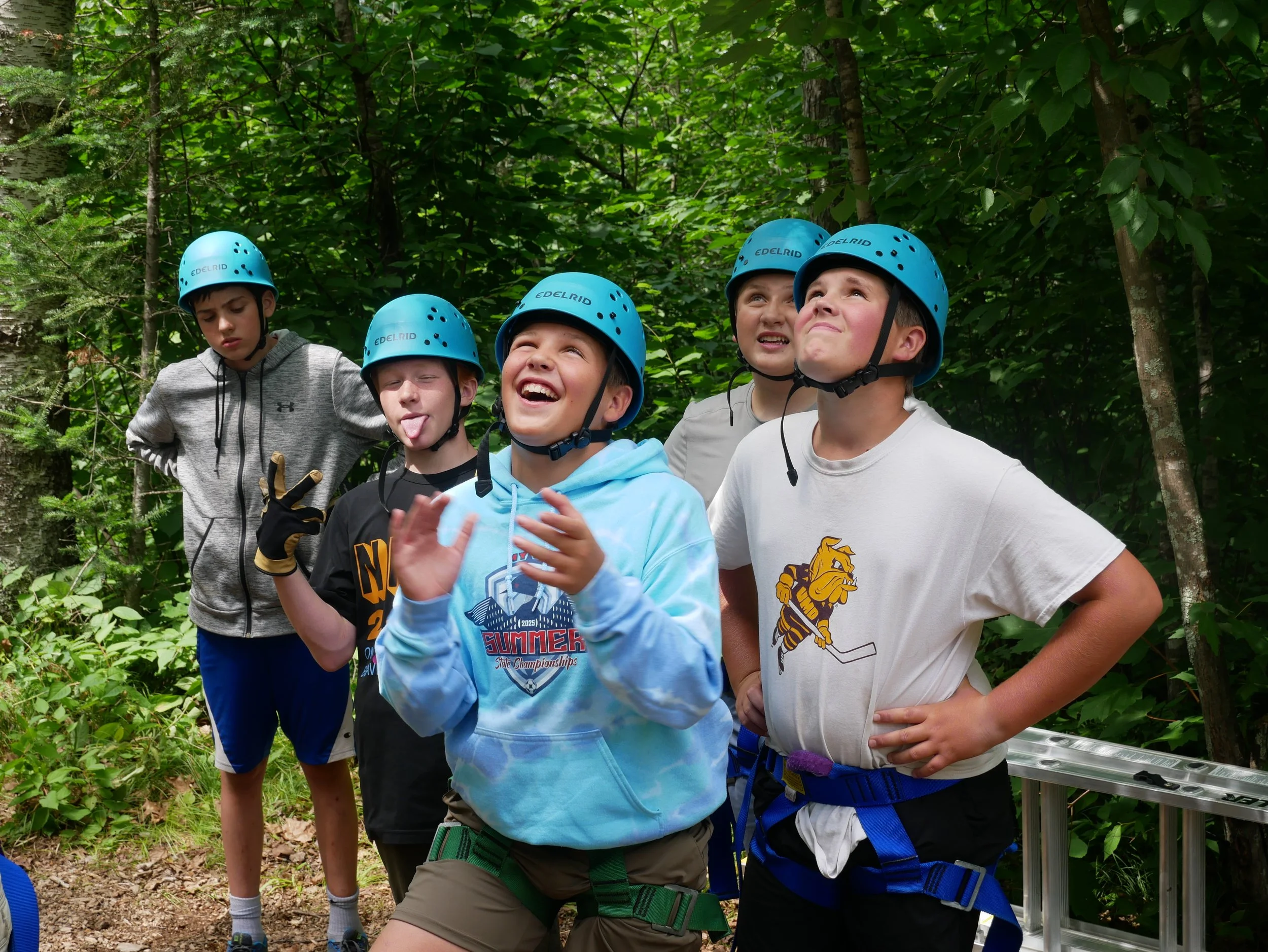 Group of five children wearing blue helmets and harnesses, standing in a wooded area, with some making playful facial expressions and gestures.
