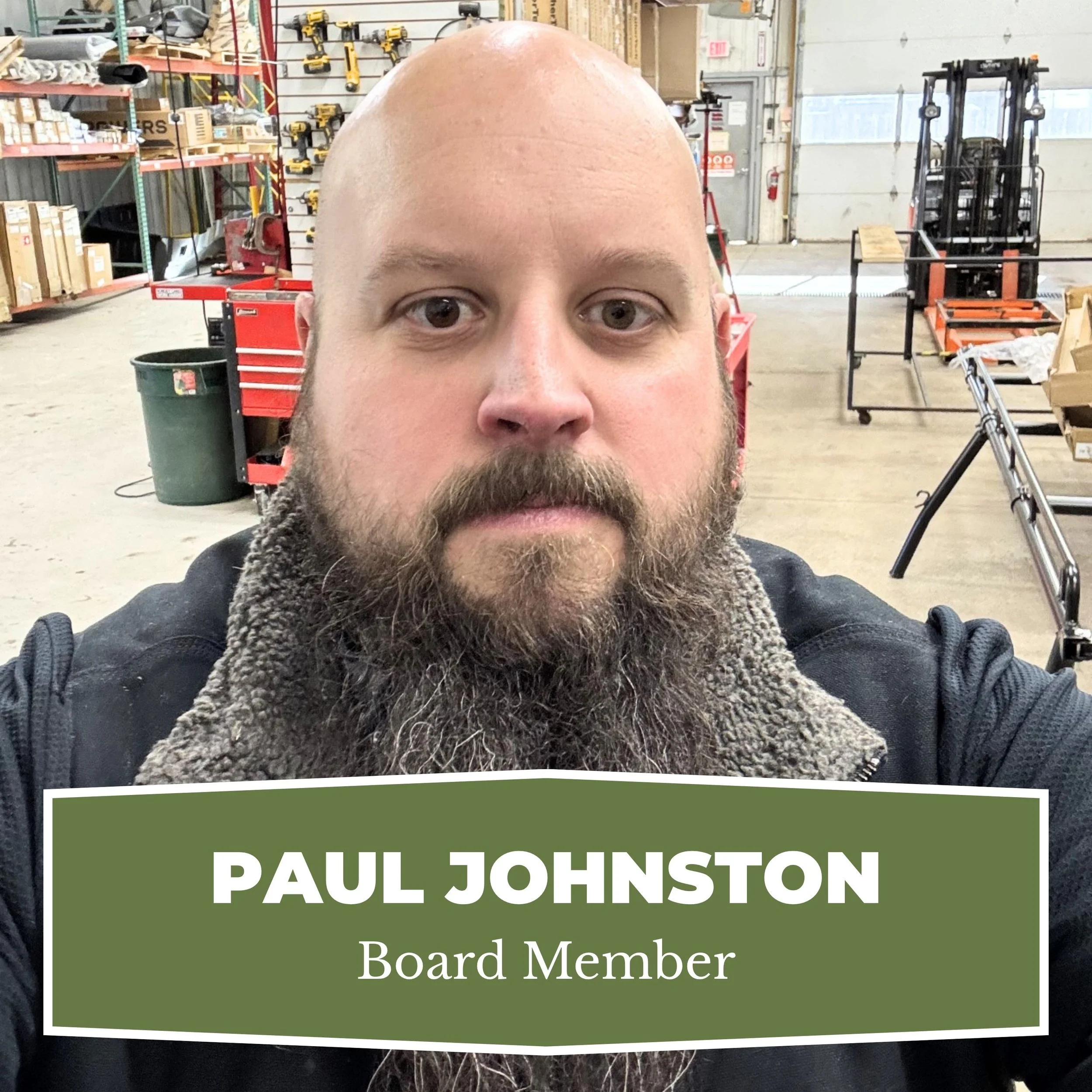 A man with a beard and a shaved head taking a selfie in a workshop or warehouse with tools and equipment in the background. A nameplate at the bottom reads 'Paul Johnston, Board Member.'
