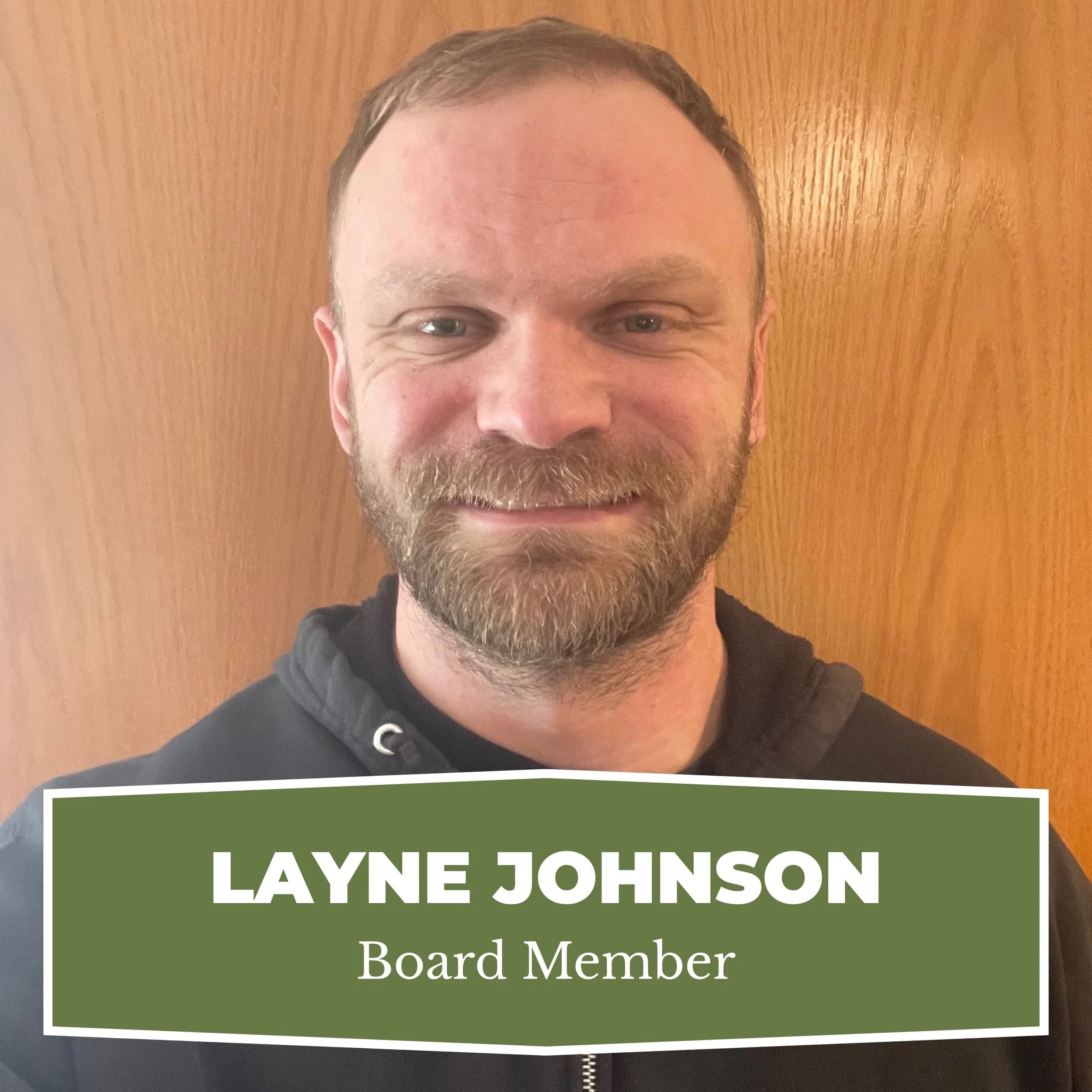A man with a beard and short hair smiling in front of a wooden background. He is wearing a dark hoodie. Overlaid on the lower part of the image is a green banner with white text that reads 'LAYNE JOHNSON' and 'Board Member'.