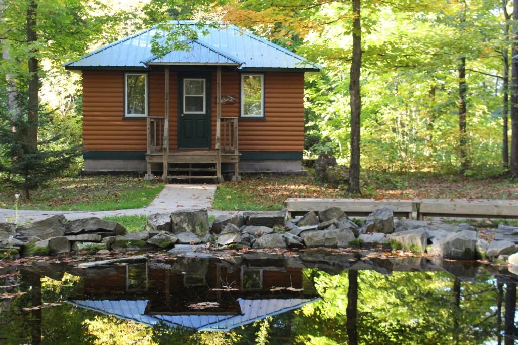 A small wooden cabin with a blue metal roof located by a pond in a forest, with trees and rocks surrounding the pond.