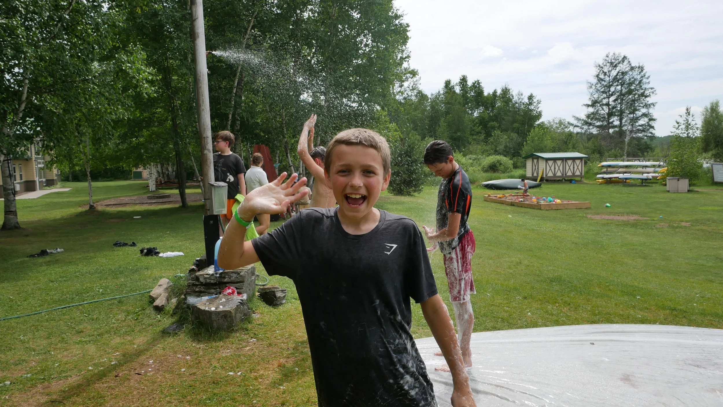 Children playing and laughing outside on a grassy area, with one boy in the foreground waving and smiling, while others are engaging in water activities.