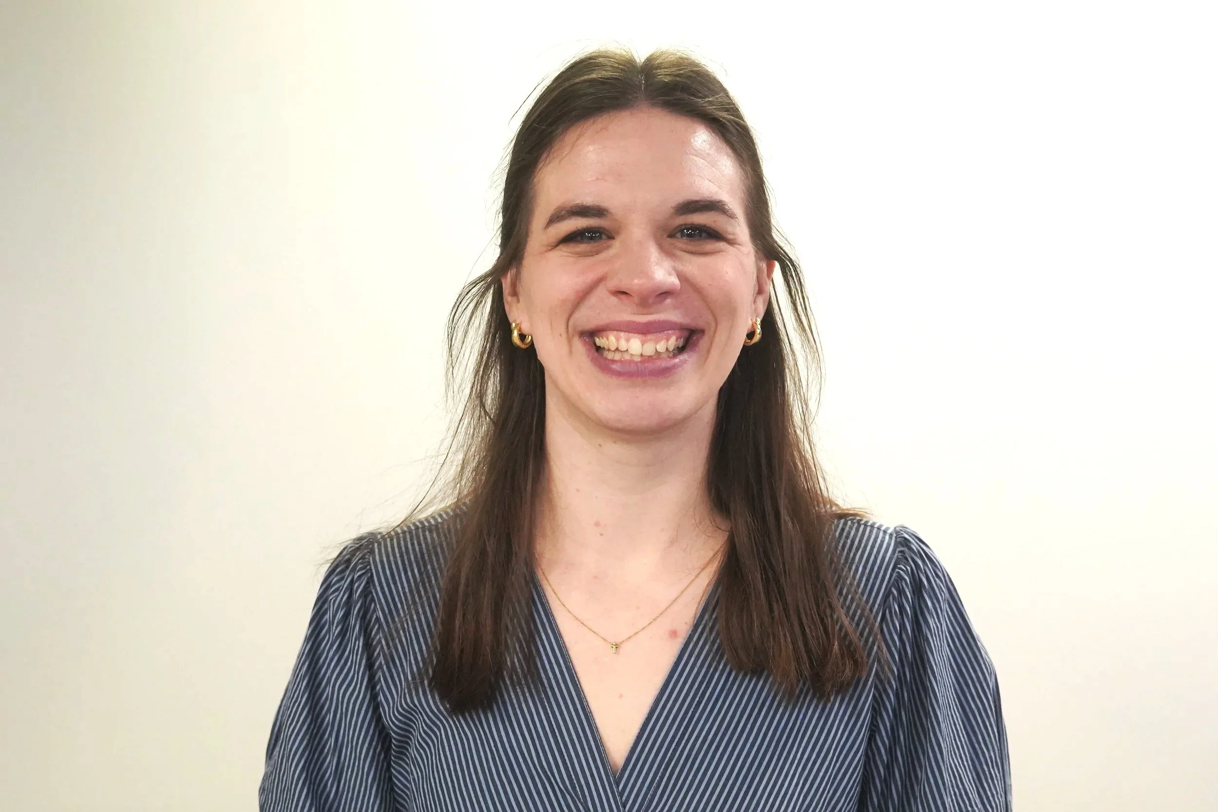 A woman with brown hair, wearing gold hoop earrings, a delicate gold necklace, and a blue striped blouse, smiling in front of a plain white wall.