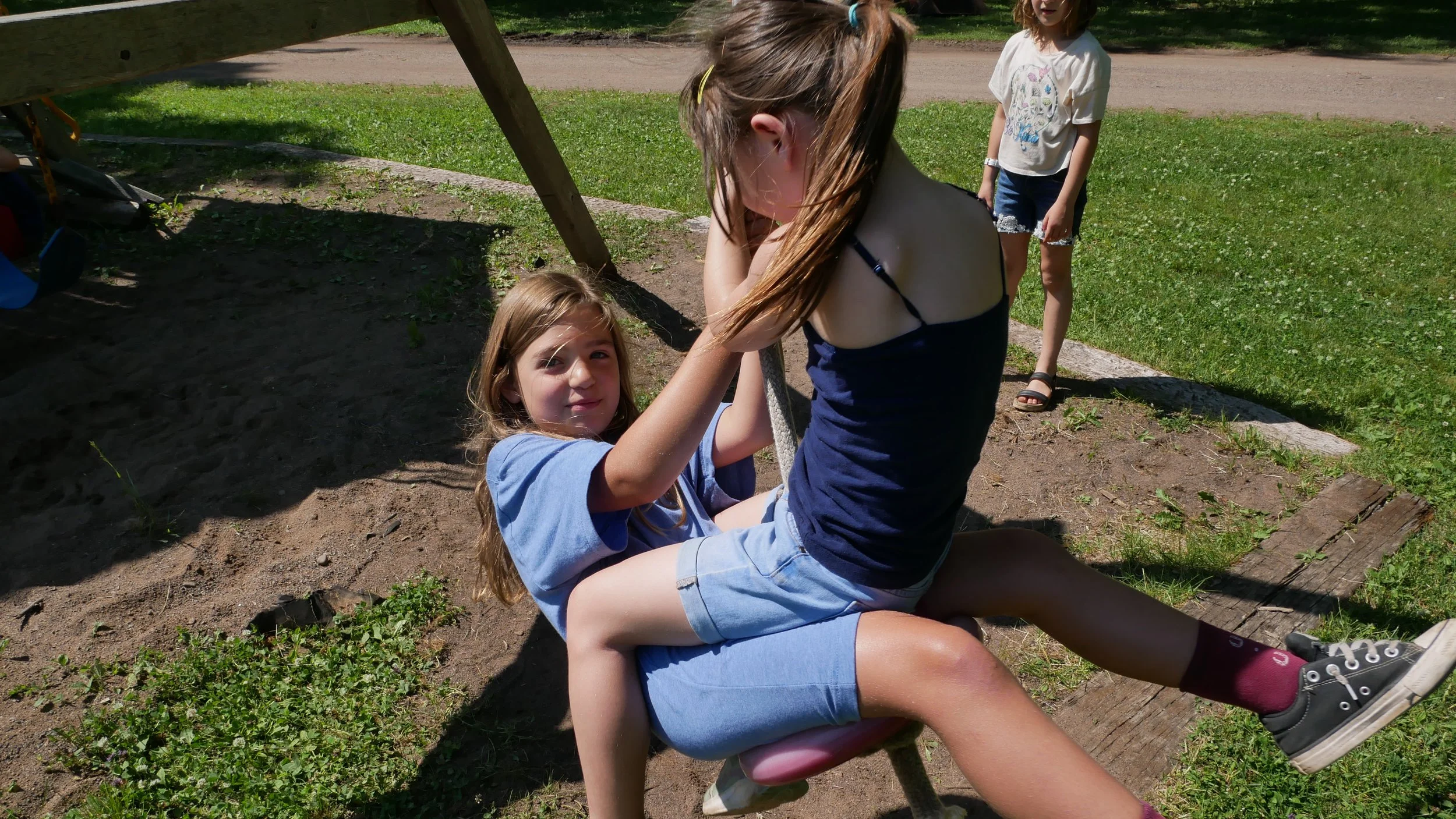 Two young girls and one older girl at a playground. The older girl is sitting on a wooden seat while the younger girl is sitting on her lap, holding onto a rope, playing on the swing set. Another girl is standing in the background on the grass. It is a sunny day with green grass and a dirt pathway in the background.