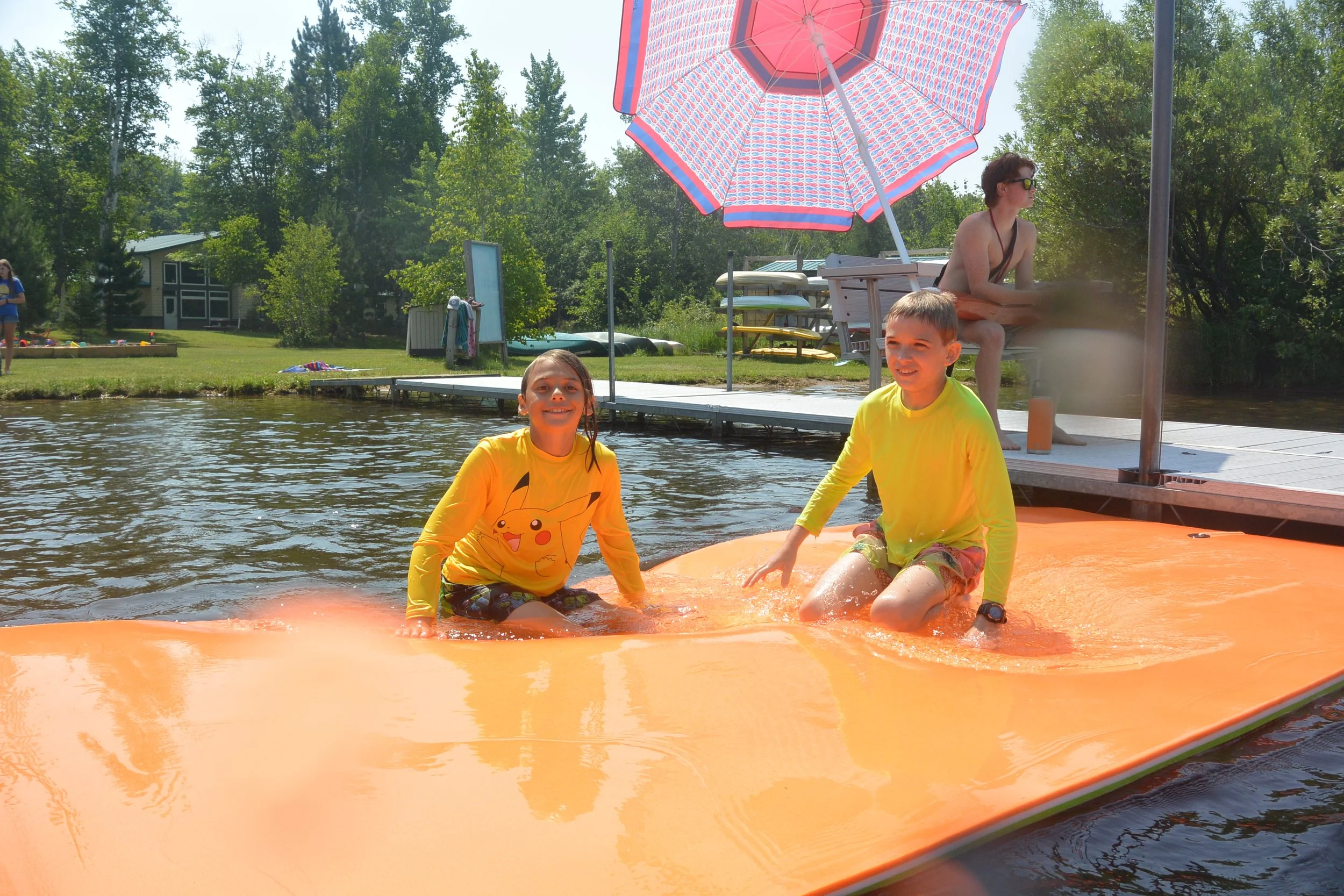 Two children in yellow long sleeve shirts are sitting on an orange float in a lake, smiling at the camera. A woman is sitting on a dock under a large pink and white striped umbrella in the background.