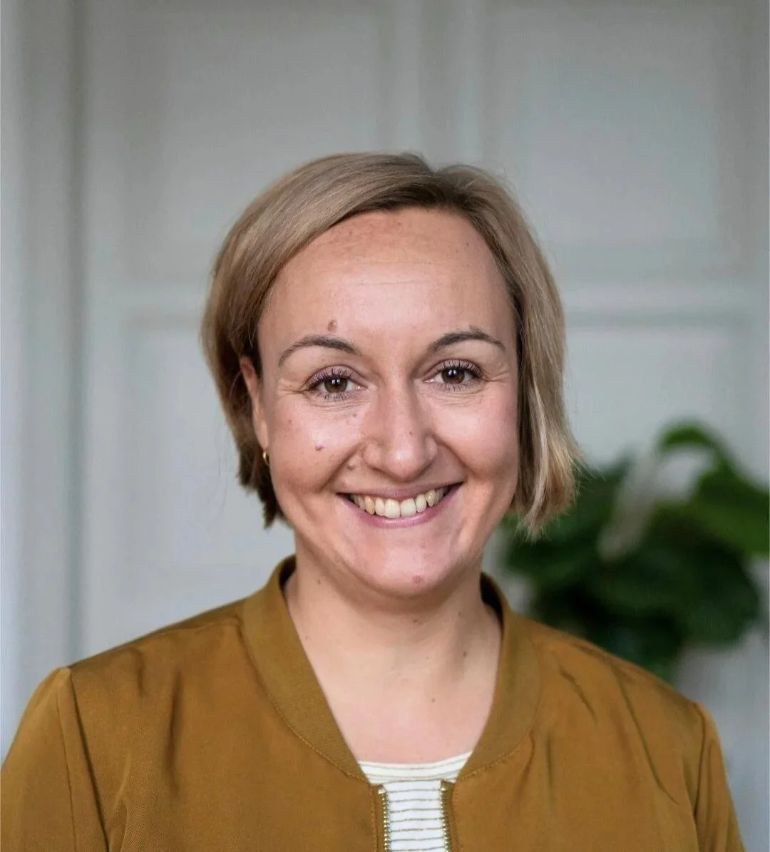 Porträt einer lachenden Frau mit kurzen, blonden Haaren, braunem Oberteil und einem weißen und braunen Hemd im Hintergrund, vermutlich in einer Innenraumumgebung.