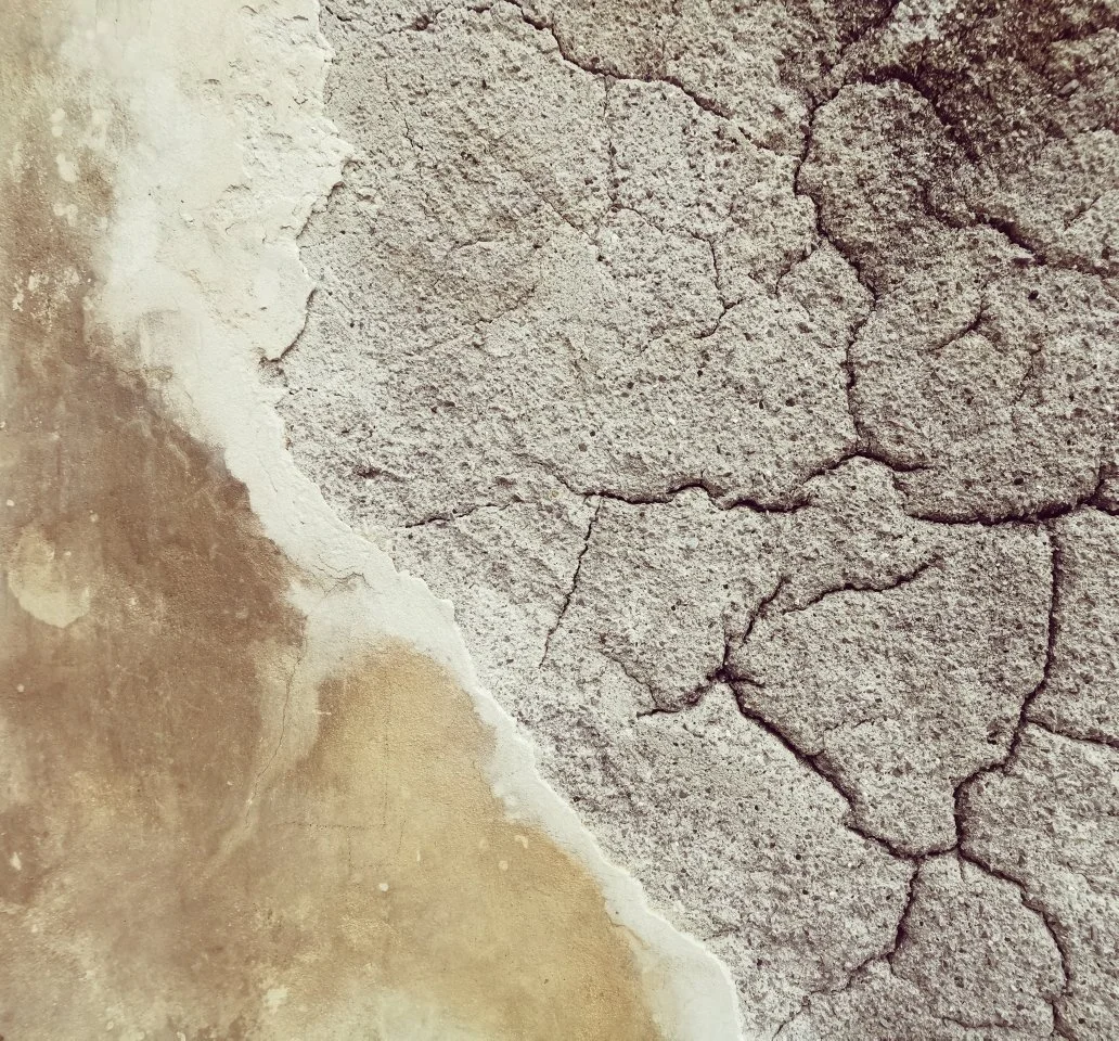 Eine Nahaufnahme einer Wand, bei der die oberste Schicht abblättert und Risse offenbart, die unteren Bereiche zeigen Staub und Verschmutzung