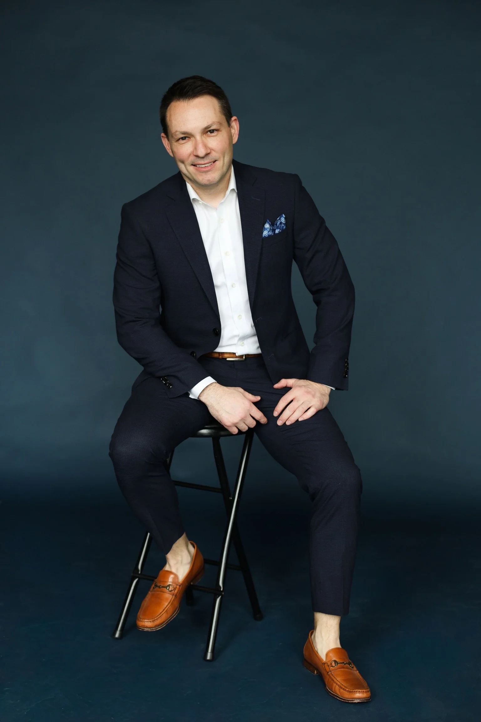 A man dressed in a navy blue blazer, white shirt, and dark pants, sitting on a stool against a dark background. He has short dark hair and is smiling slightly. He is wearing brown loafers and a blue patterned pocket square.