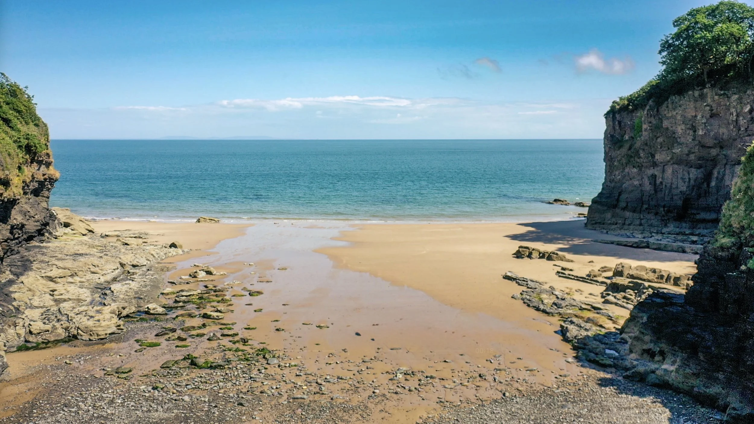 A secluded beach with sandy shore, rocky cliffs on both sides, green vegetation on the cliffs, blue skies with a few clouds, and calm ocean water.