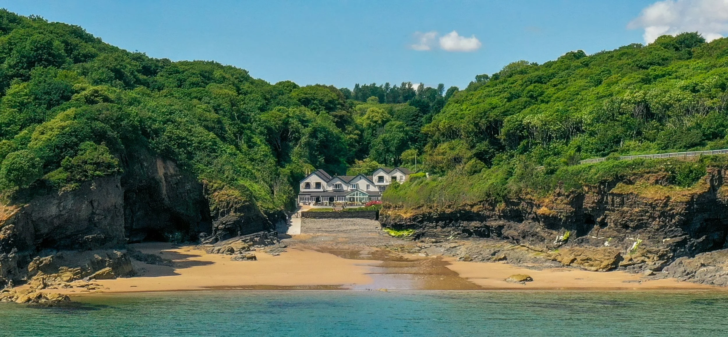A secluded beach with cliffs and lush green hills surrounding a large house with multiple levels, nestled on the hillside. The sandy shore meets turquoise water, with a partly cloudy sky overhead.
