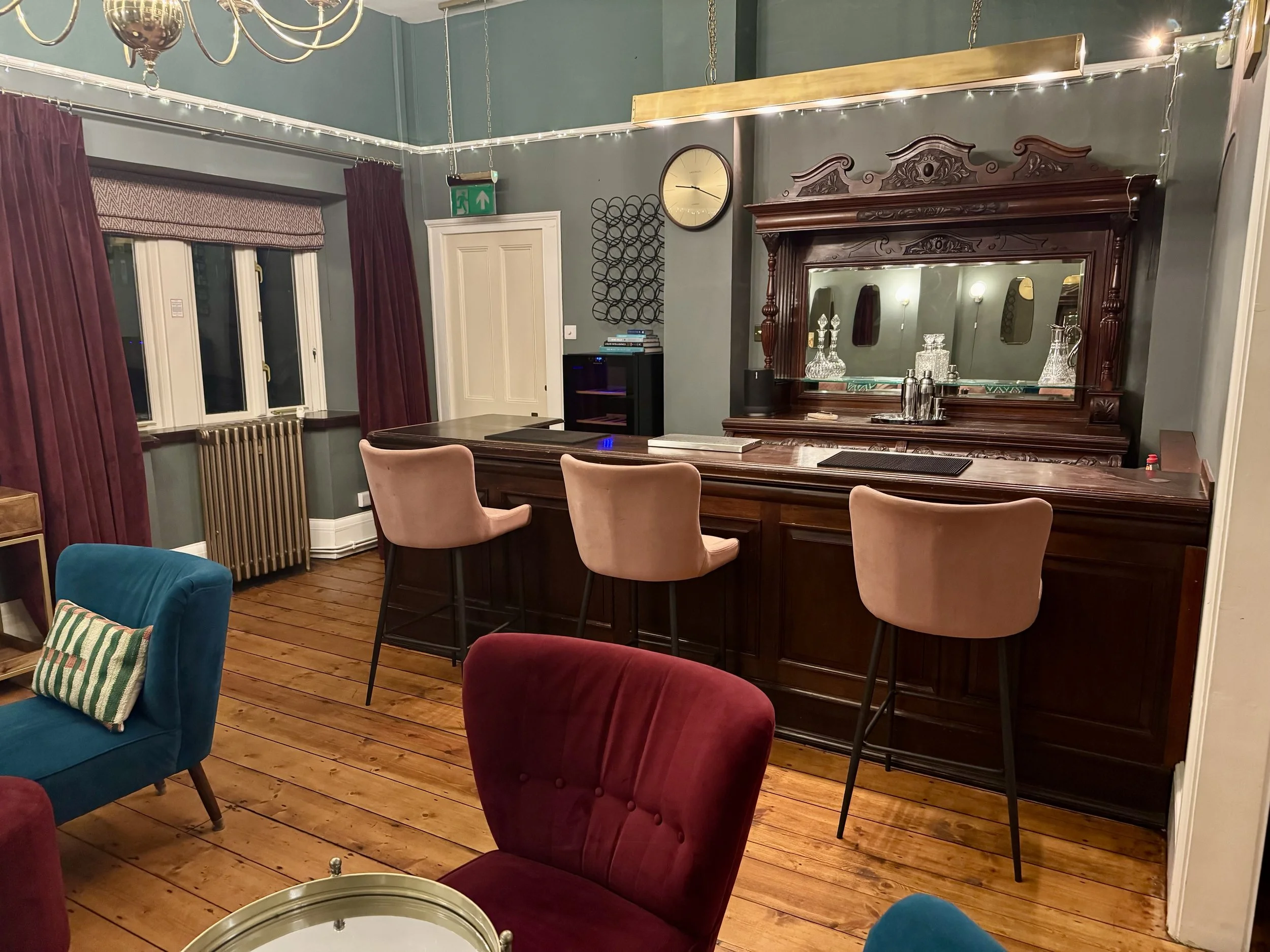 A cozy entertainment room with hardwood floors, featuring a large ornate wooden mirror, pink upholstered chairs, a blue armchair with cushions, a black cabinet, and a window with red curtains and a patterned valance, decorated with string lights, a clock, and wall art.
