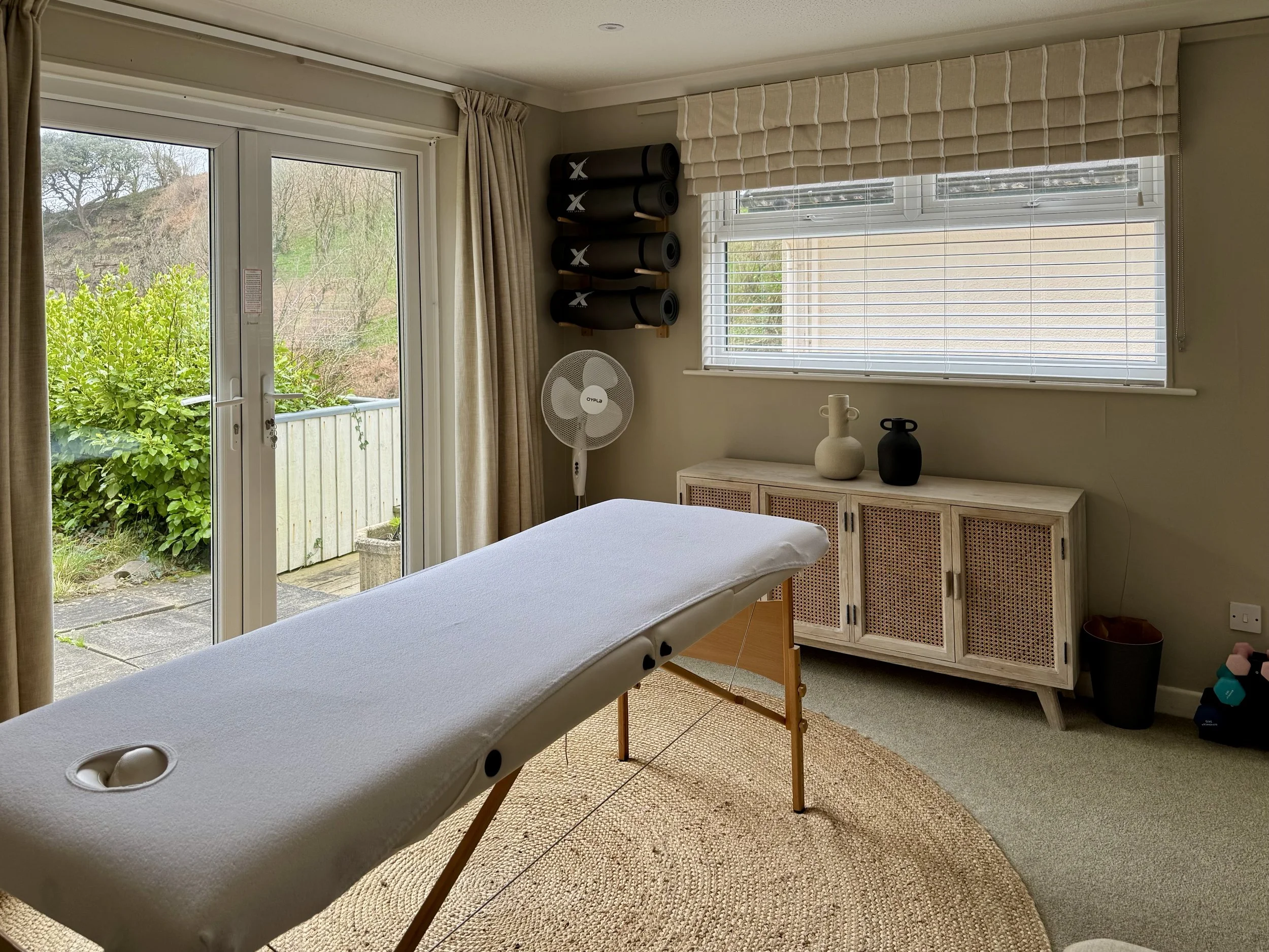 Interior of a room with a massage table, a sideboard with decorative vases, a window with blinds, and a wall-mounted rack holding four black yoga mats, adjacent to a door leading outside to a garden.