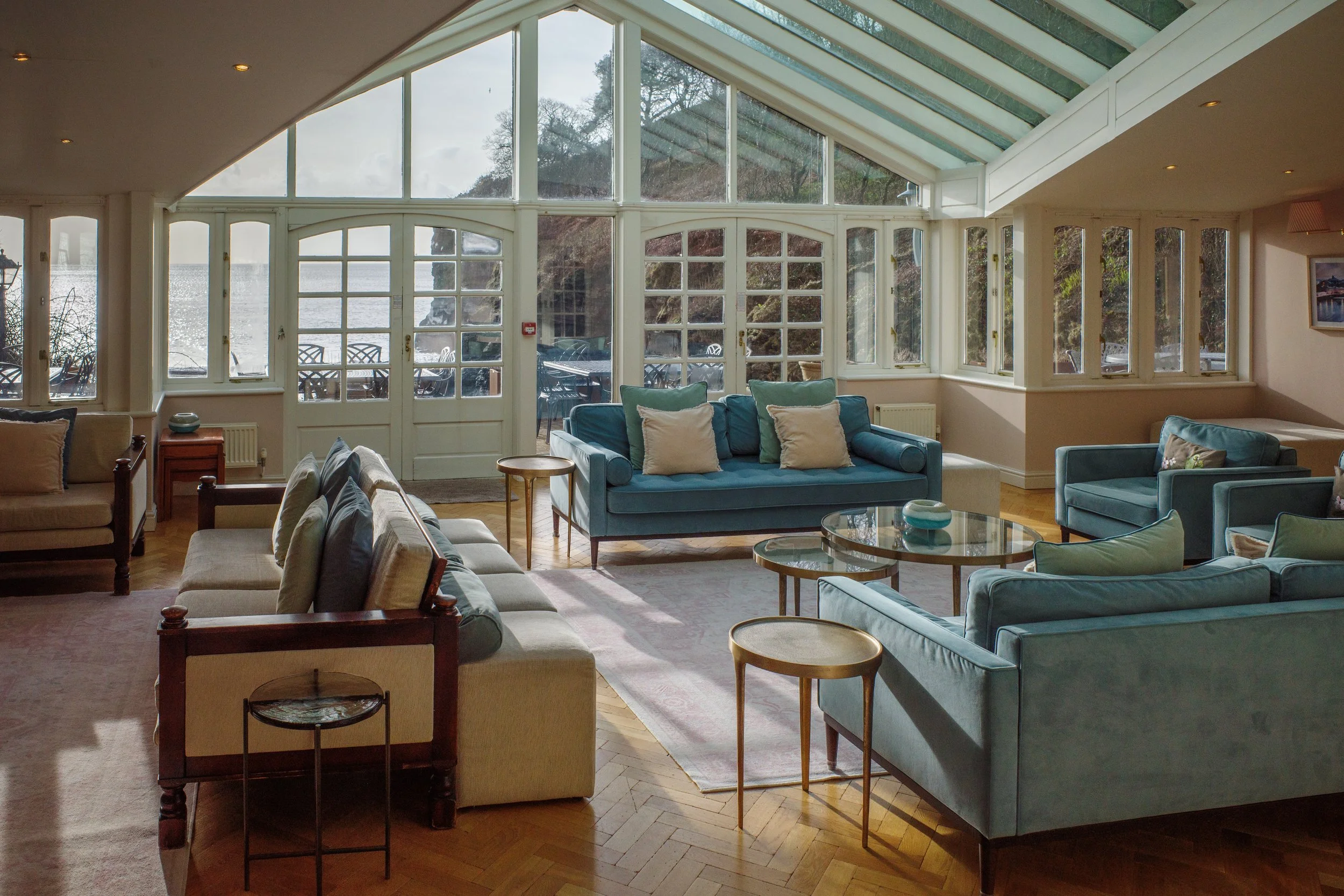 Living room with large windows and a view of the outdoors, furnished with various sofas, armchairs, side tables, and a coffee table, with a sloped glass ceiling.