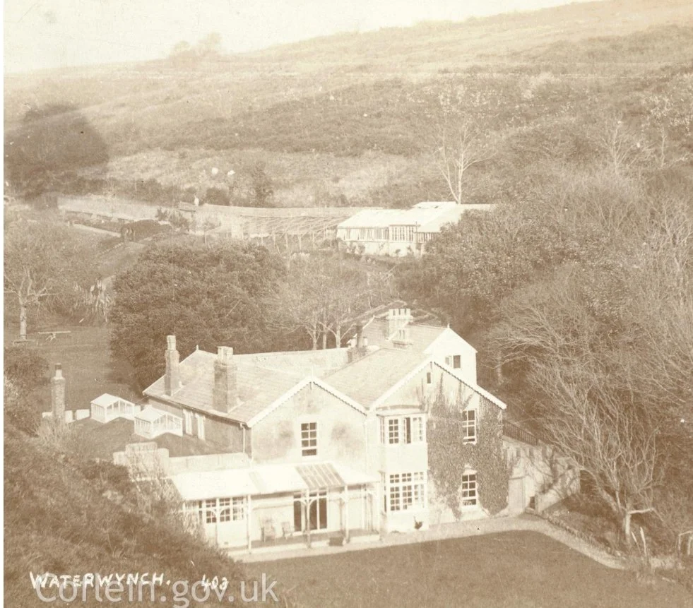 A sepia-toned photo of a large house with chimneys, surrounded by trees, with a rural landscape in the background.