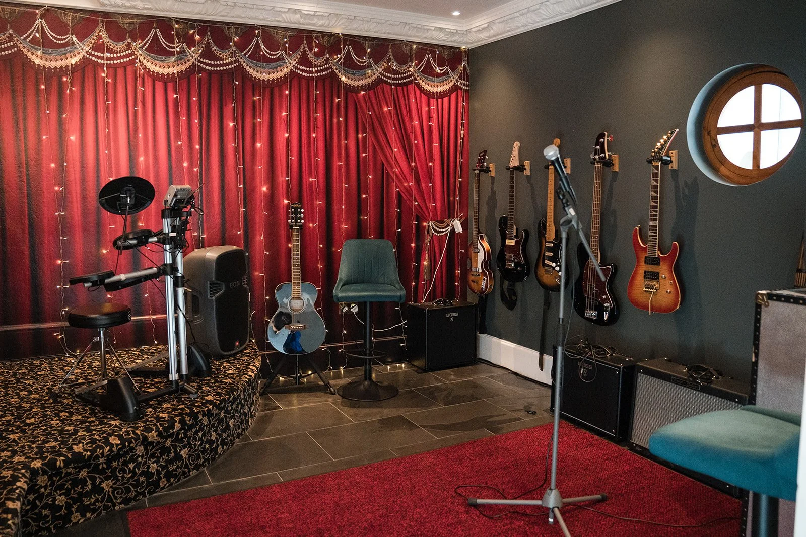 A music rehearsal room with red velvet curtains, string lights, five guitars on the wall, a microphone stand, an electric drum pad, and a chair in the center.