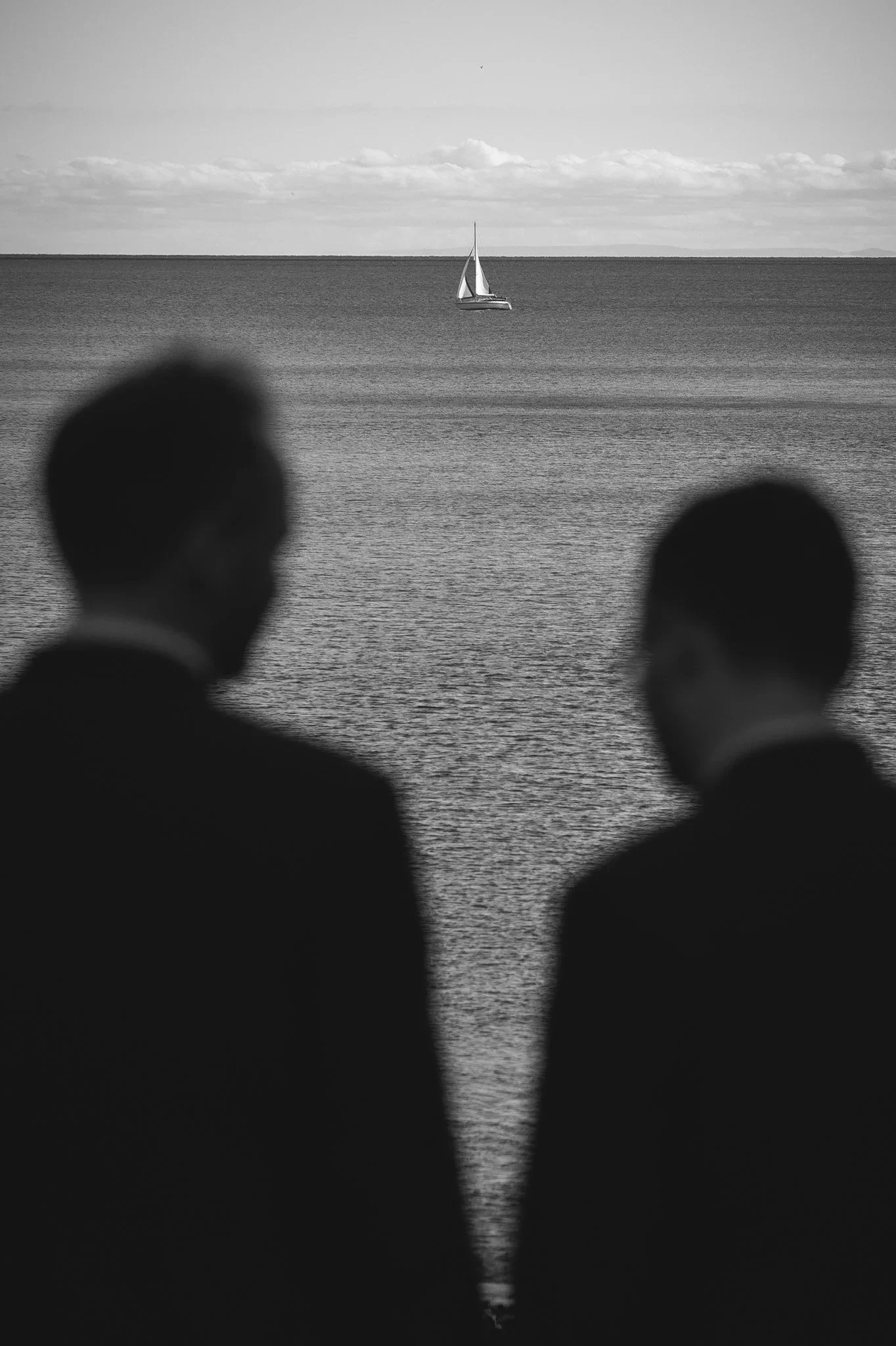 Black and white photo of two blurred figures in formal attire in the foreground, looking at a sailboat on the water in the distance.