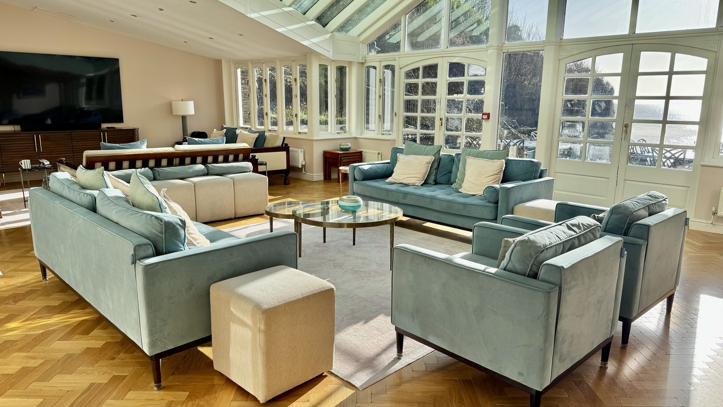 Bright sunlit living room with pastel blue and beige sofas, a glass-top coffee table, and large windows with a view of outdoor furniture and water. Panoramic sea views.