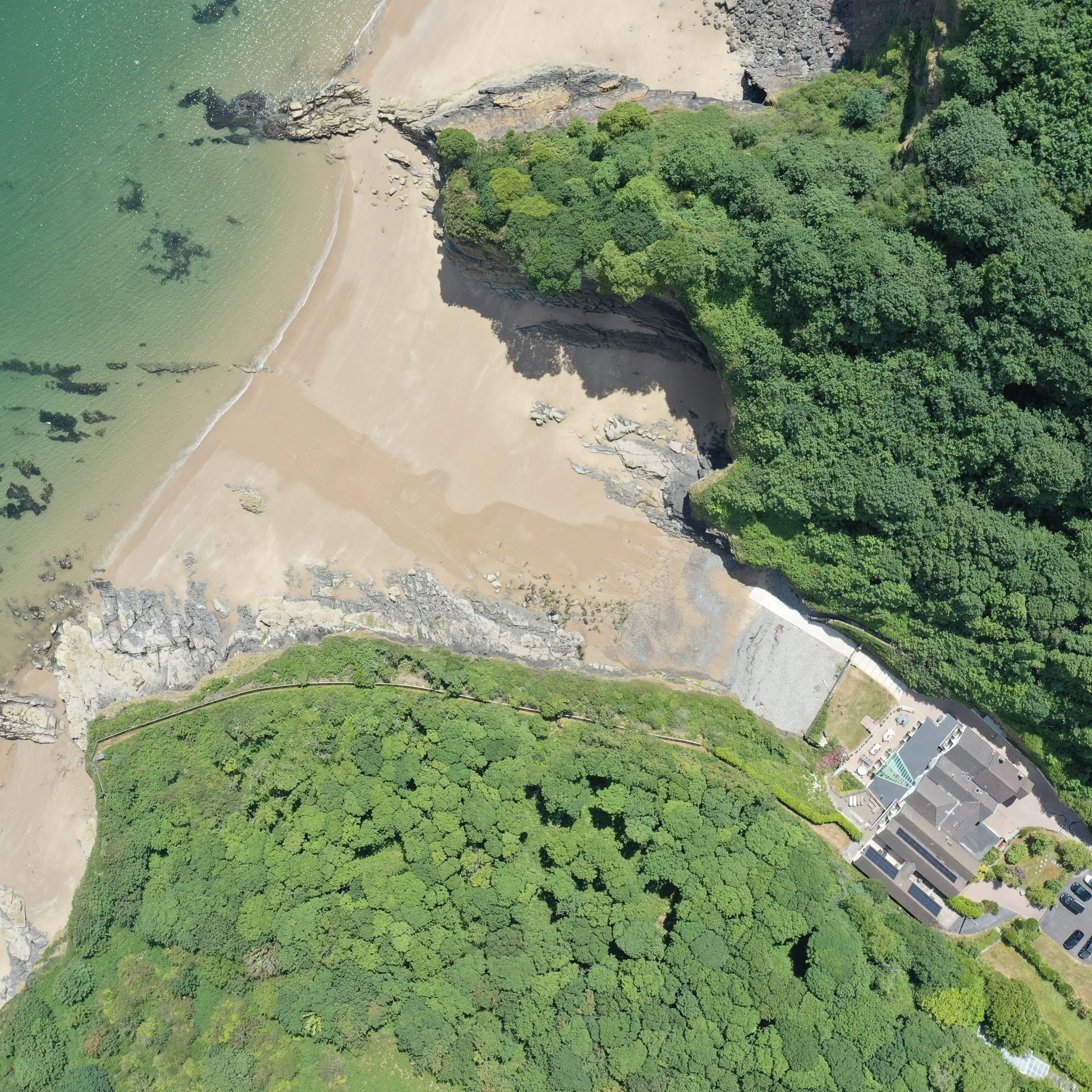An aerial view of a beach with green water, a sandy shoreline, cliffs, and lush green trees. There are also some buildings and parked cars visible.