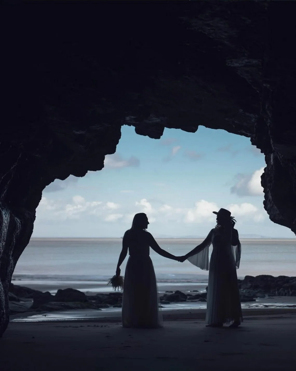 Silhouette of two women holding hands on a beach viewed from inside a rocky cave, with ocean and sky in the background.