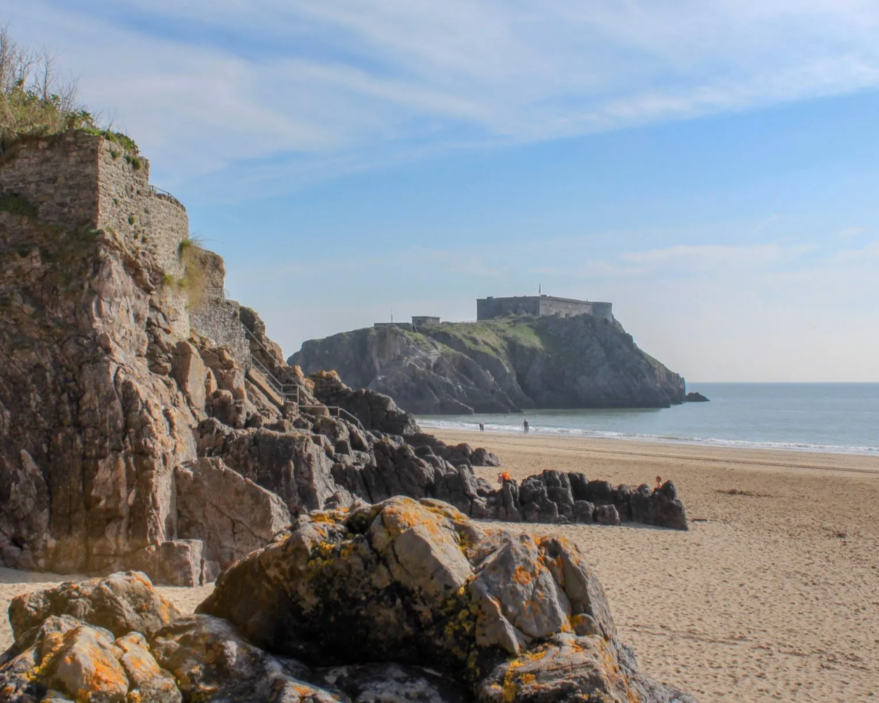 Rocky beach with stairs leading up a cliff to a historic fortress on a hill, blue sky with light clouds, few people walking on the sandy shore.