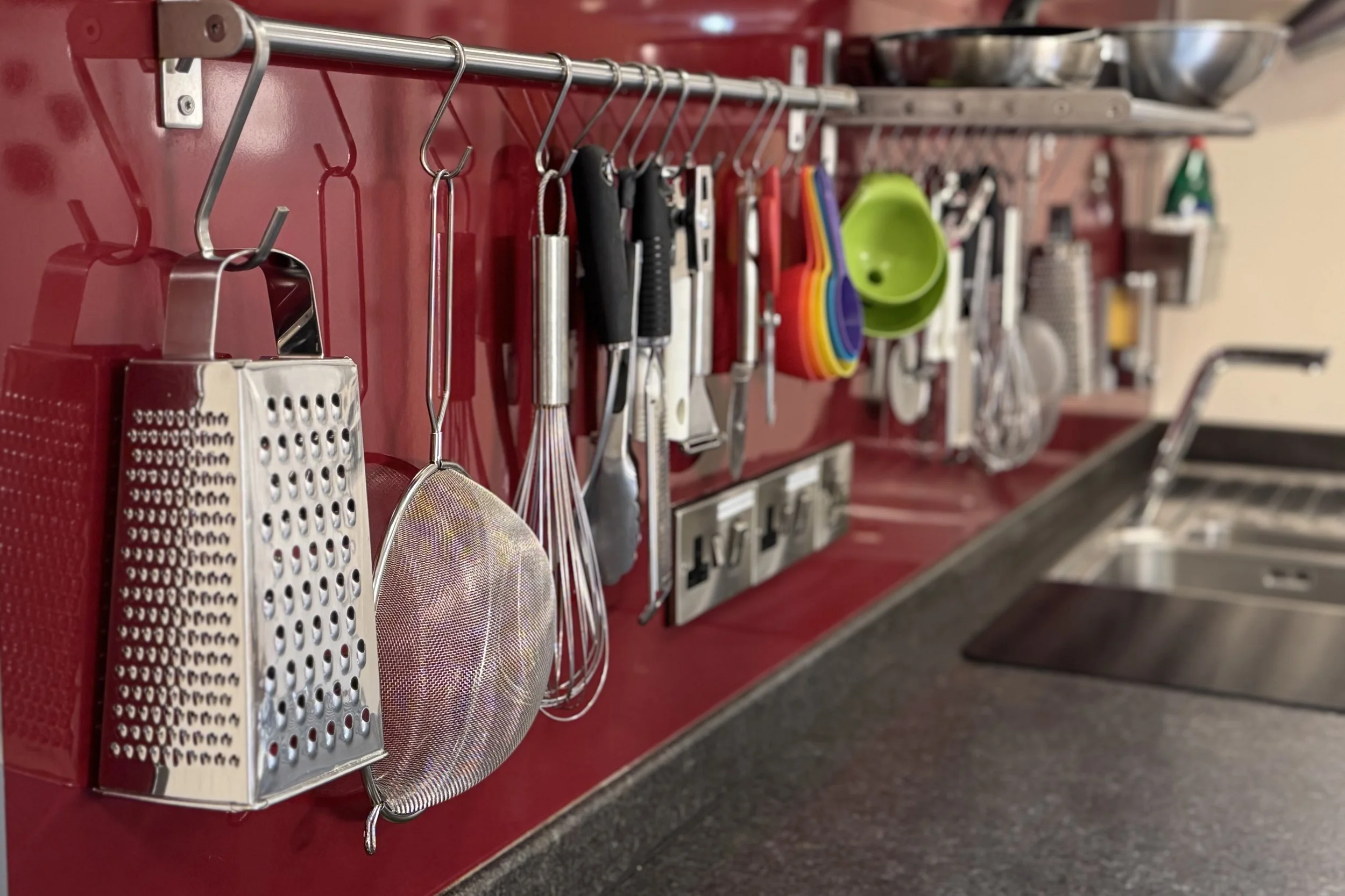 Kitchen utensil storage with grater, whisk, tongs, and measuring cups hanging on a red wall-mounted rack inside a kitchen.