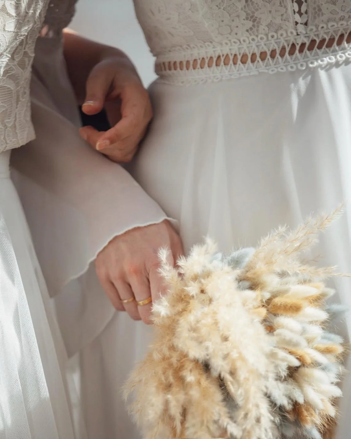 Close-up of a woman in a white lace dress holding a bouquet of dried flowers, with her other hand displaying a gold ring.