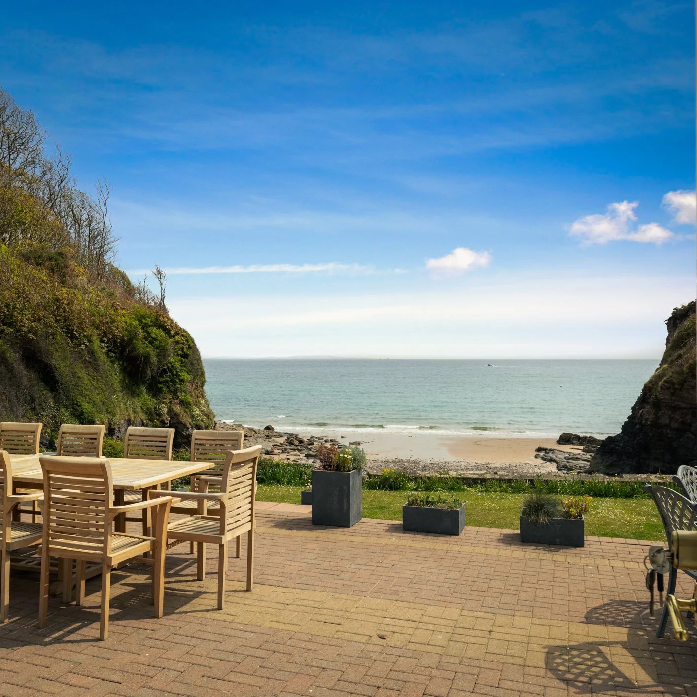 Ocean view with sandy beach framed by cliffs on either side, outdoor wooden table and chairs on a patio, potted plants, and a clear blue sky.