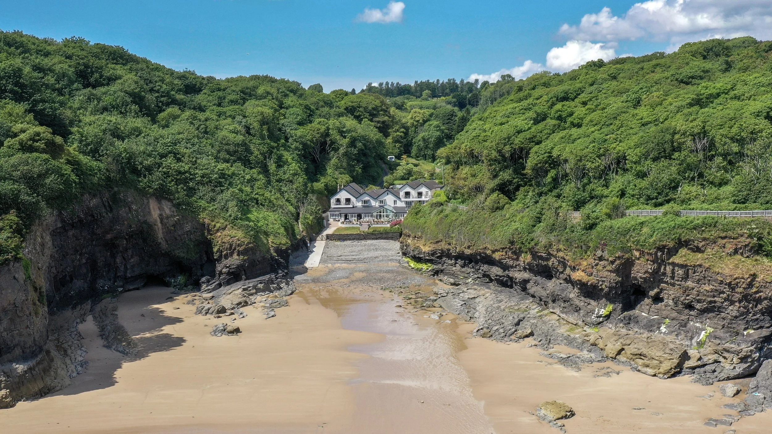 A beach with sandy shore and rocky cliffs on both sides, with a house on a hill overlooking the beach and green hills in the background under a blue sky.