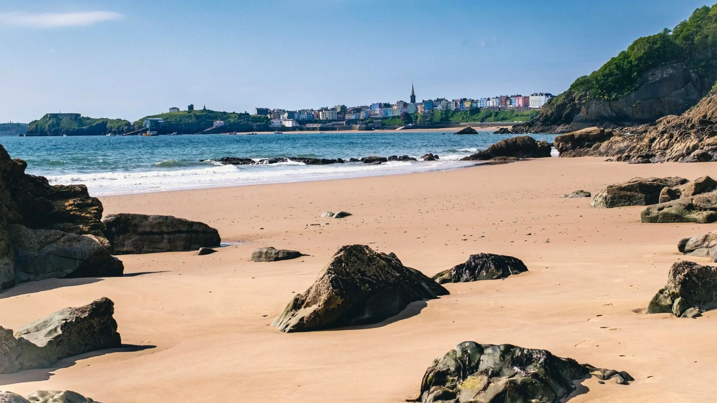 Sandy beach with rocks along the shore, calm ocean water, colorful buildings on a hill in the background, clear blue sky with a few clouds.