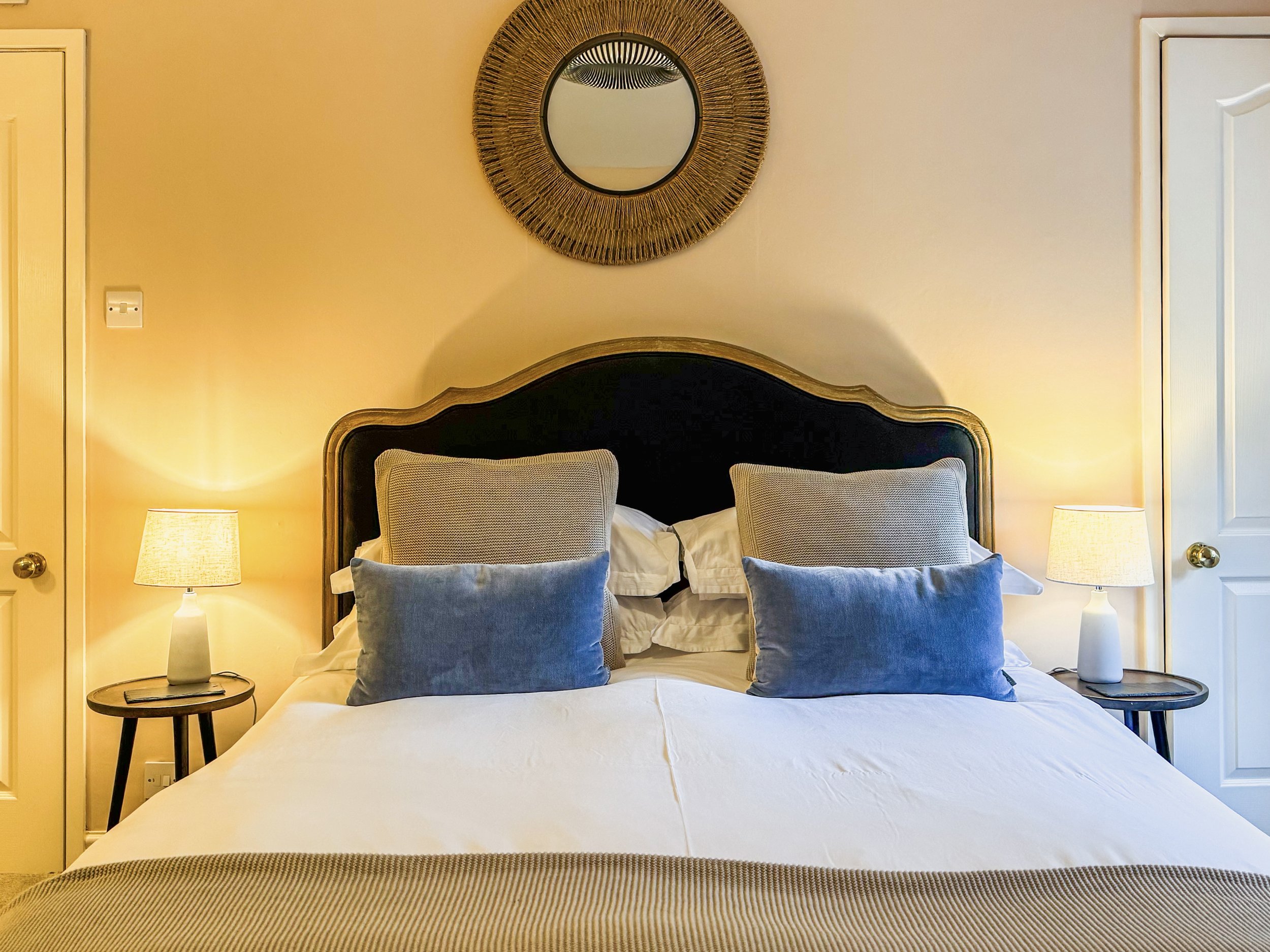 A neatly made bed with a black headboard, multiple pillows in shades of gray and cream, two bedside lamps, a round mirror with a woven frame hanging above the headboard, and two closed closet doors on either side of the bed in a cozy bedroom.