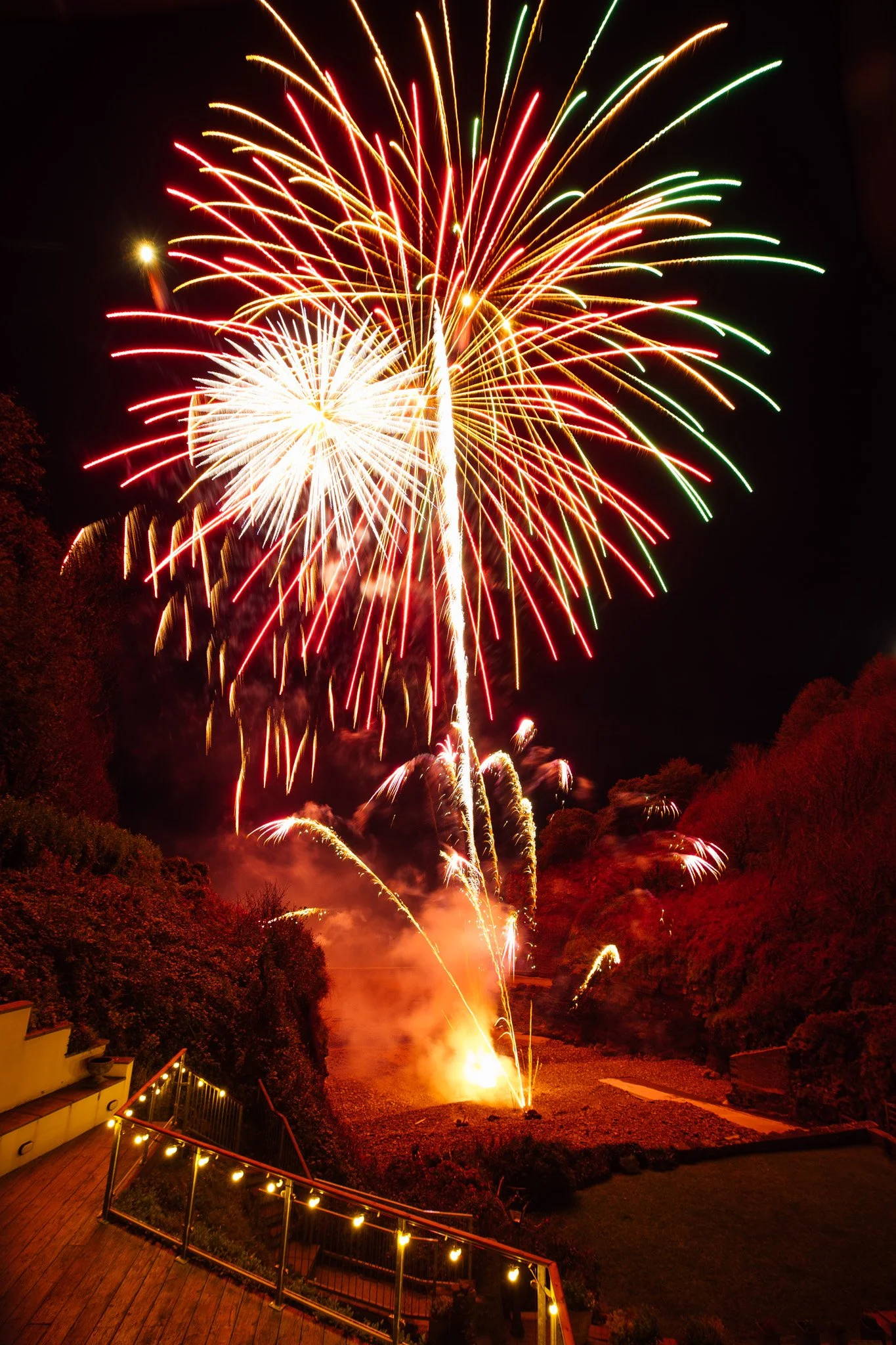 Colorful fireworks exploding in the night sky over an outdoor area with trees, a wooden deck, and string lights.