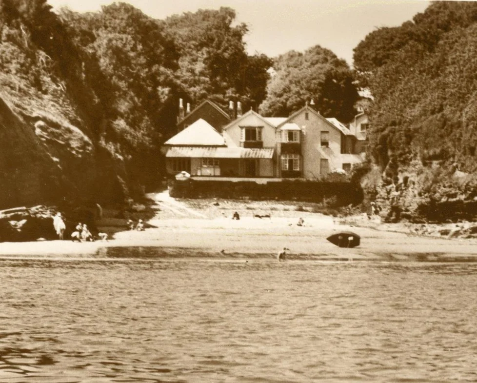 A sepia-toned photograph of a house by the water's edge, with trees surrounding the property and a beach with a few people, umbrellas, and boats in the foreground.