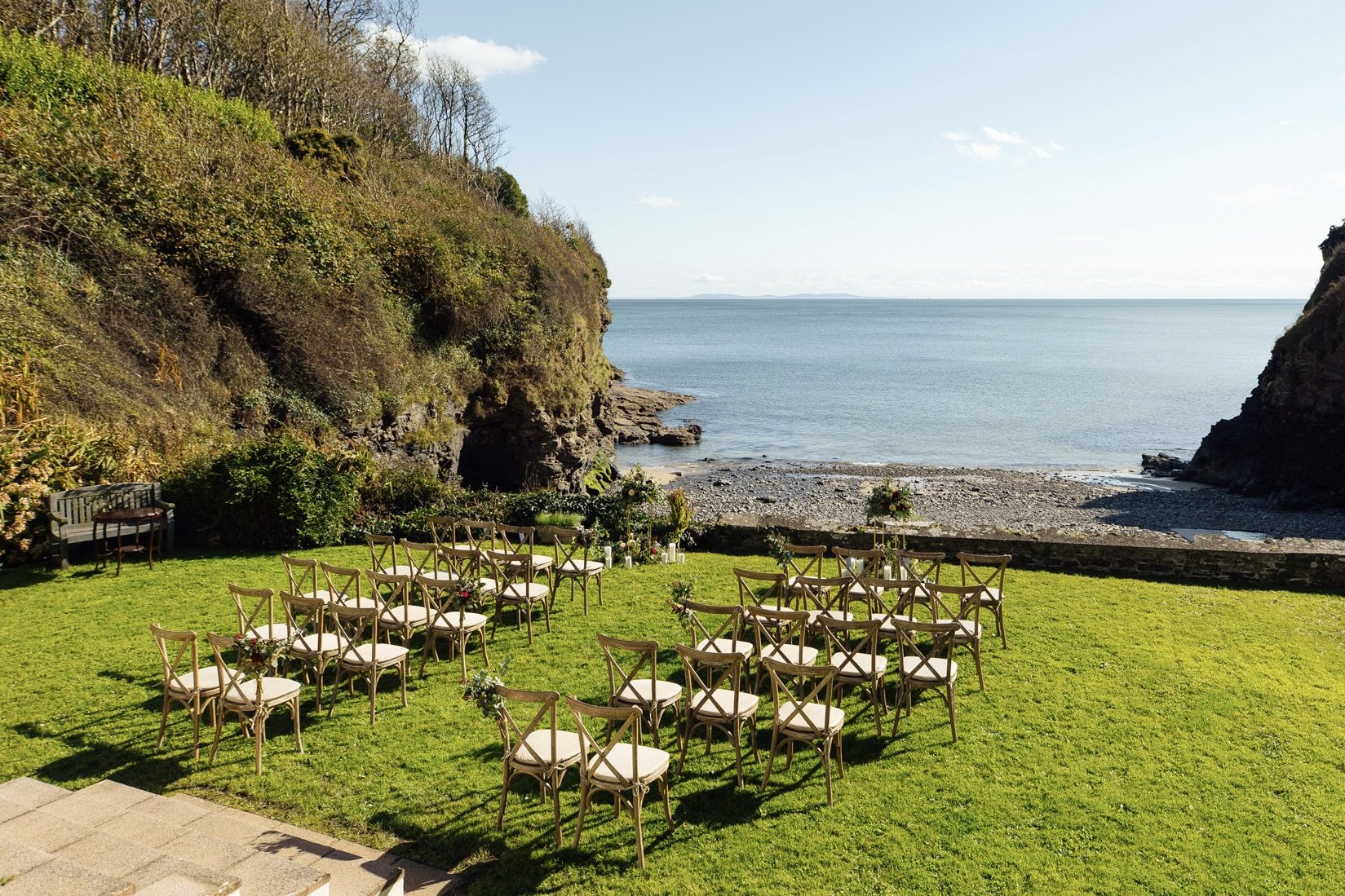 Outdoor wedding setup on a grassy lawn with chairs facing the ocean, between two rocky cliffs on a sunny day.