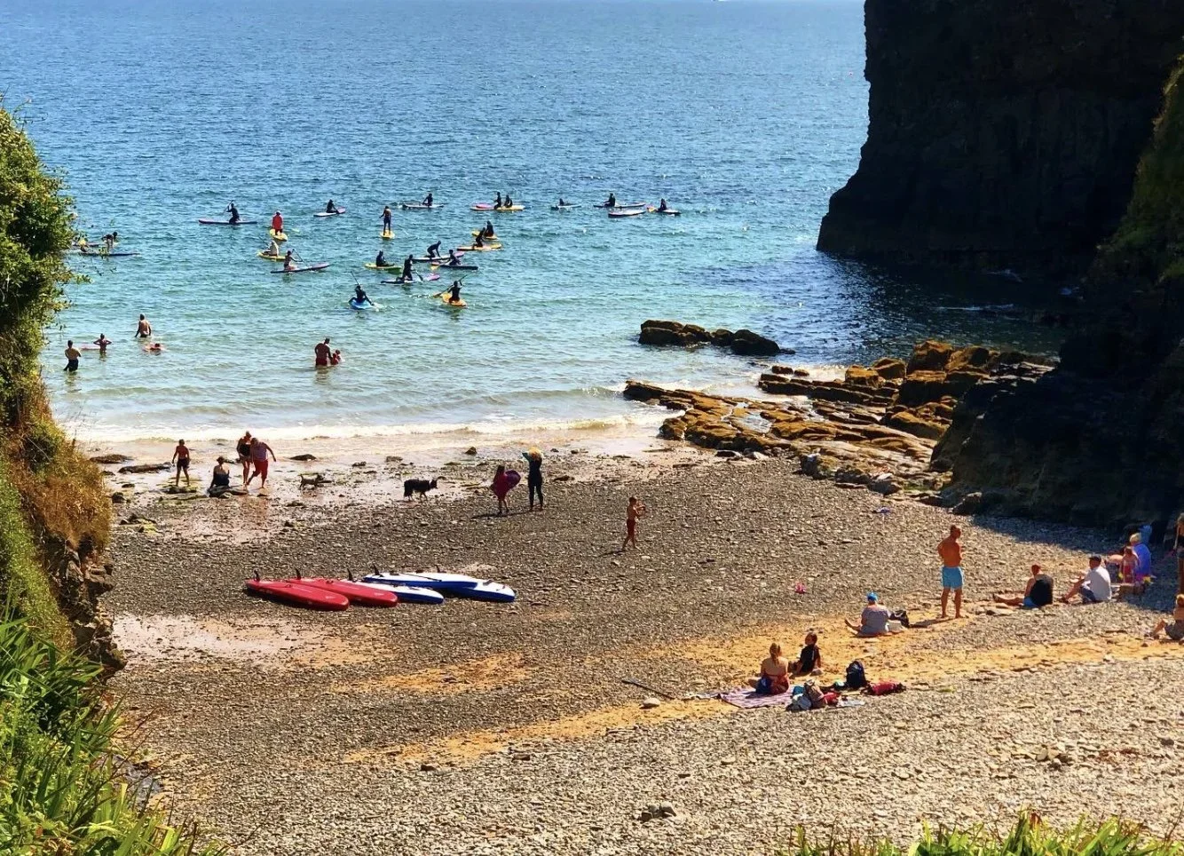 A beach scene with a rocky cliff on the right, people swimming and paddleboarding in the water, and others sitting on the pebbled shore, with several kayaks placed on the sand.