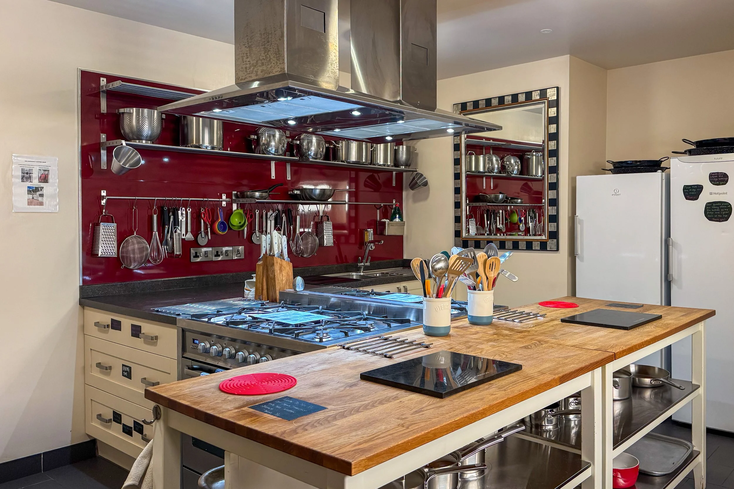 A kitchen with a red wall and stainless steel cabinetry. There are various kitchen utensils hanging on the wall, a stovetop with four burners, and a wooden countertop with two red silicone hot pads and kitchen utensils in white containers. A white refrigerator is visible in the background.