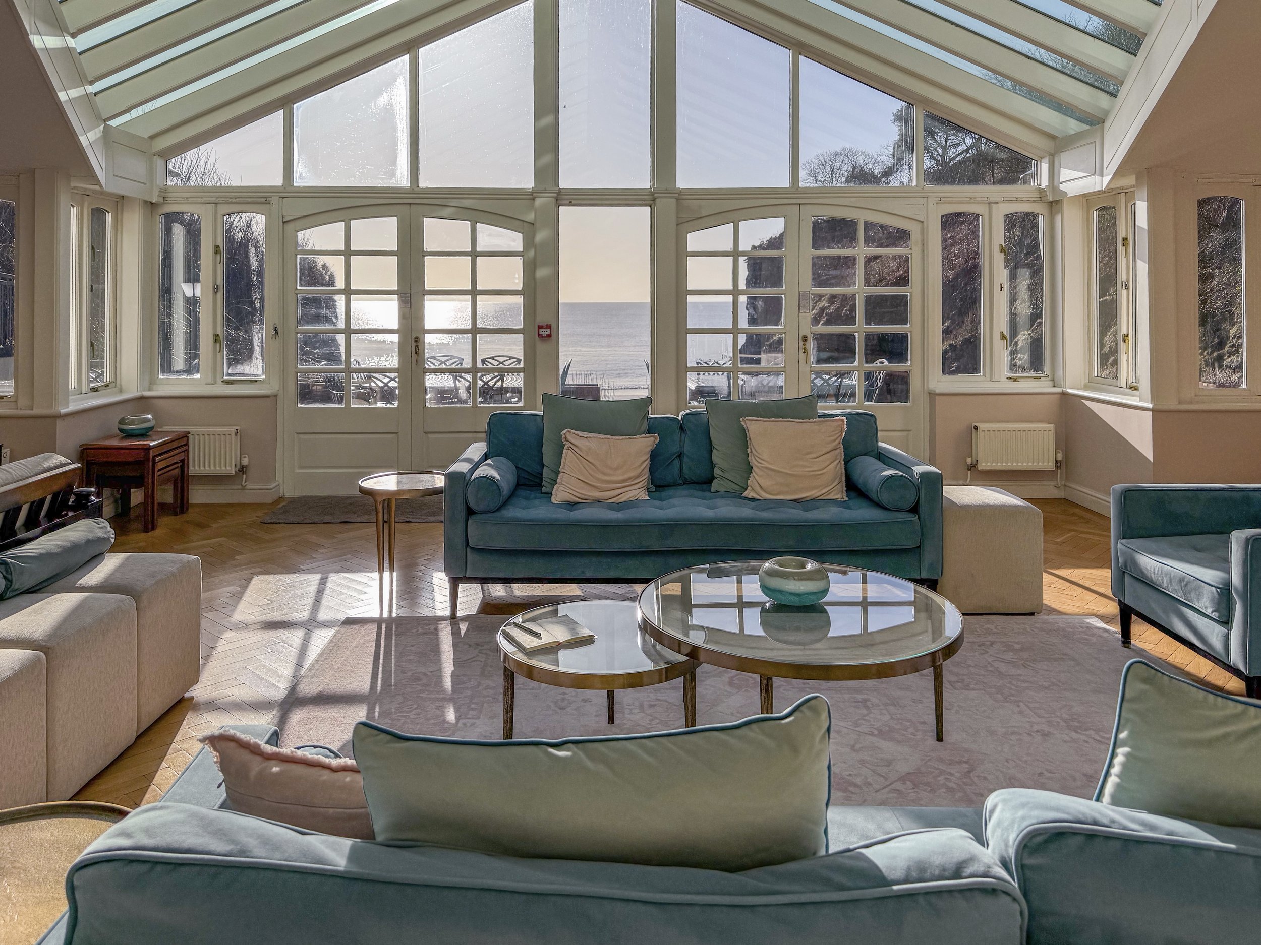 Living room with large glass windows and door overlooking the ocean, with Blue sofas and a round glass coffee table.