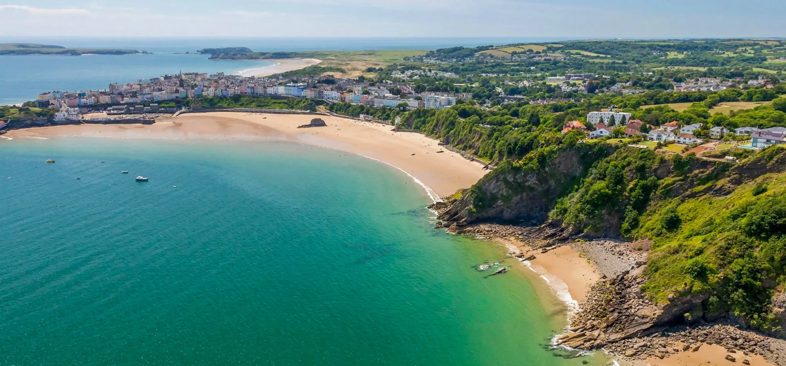 Aerial view of a coastal town with a sandy beach, turquoise water, green hillside, and colorful buildings along the shoreline.