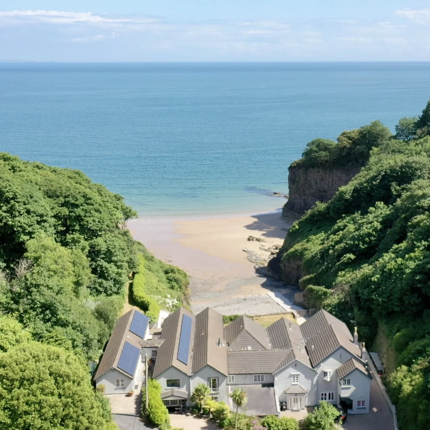 A coastal view with a sandy beach, turquoise ocean, lush green cliffs, and white houses with solar panels on rooftops.