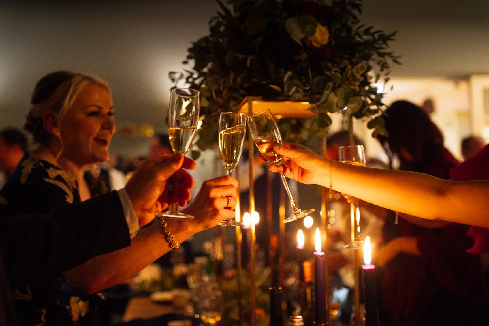 People clinking champagne glasses at a celebration with candles and candles on a table, with a woman smiling in the background.