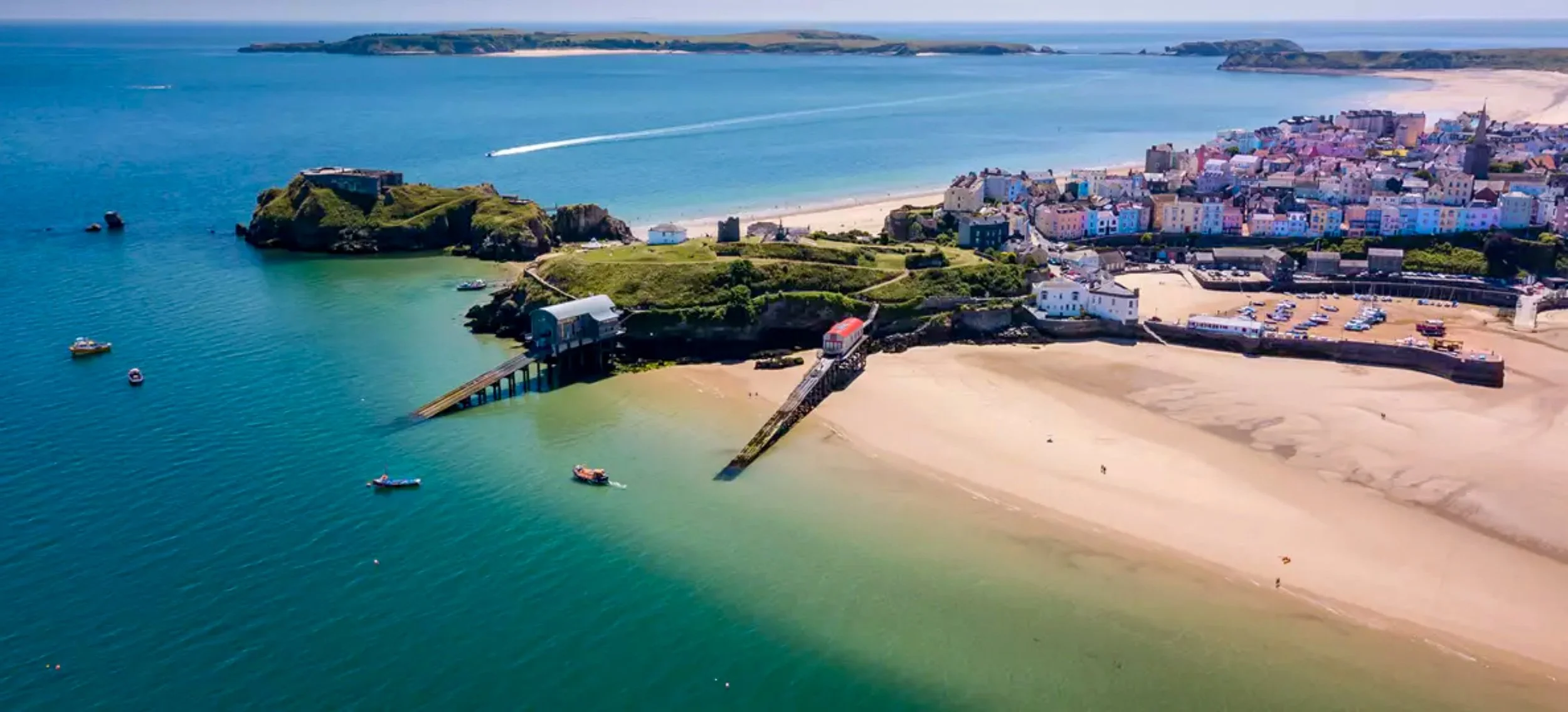Aerial view of a seaside town with a sandy beach, green hills, boats in the water, and colorful buildings near the shore.