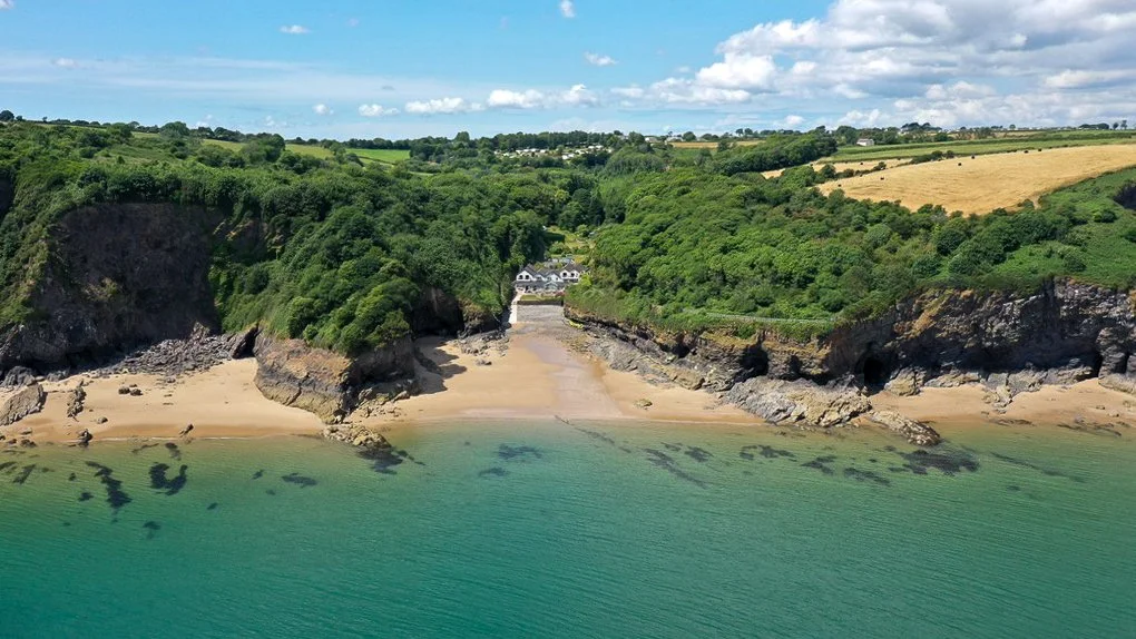 A scenic coastal landscape with a sandy beach, rocky cliffs, and lush green hills under a partly cloudy sky.