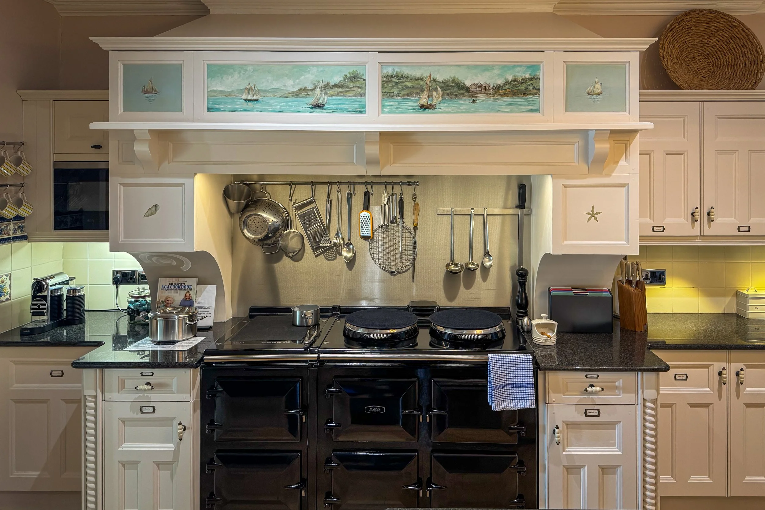 Kitchen with white cabinets, black stovetop, and various kitchen utensils hanging above counter, with a framed seaside painting on the wall.