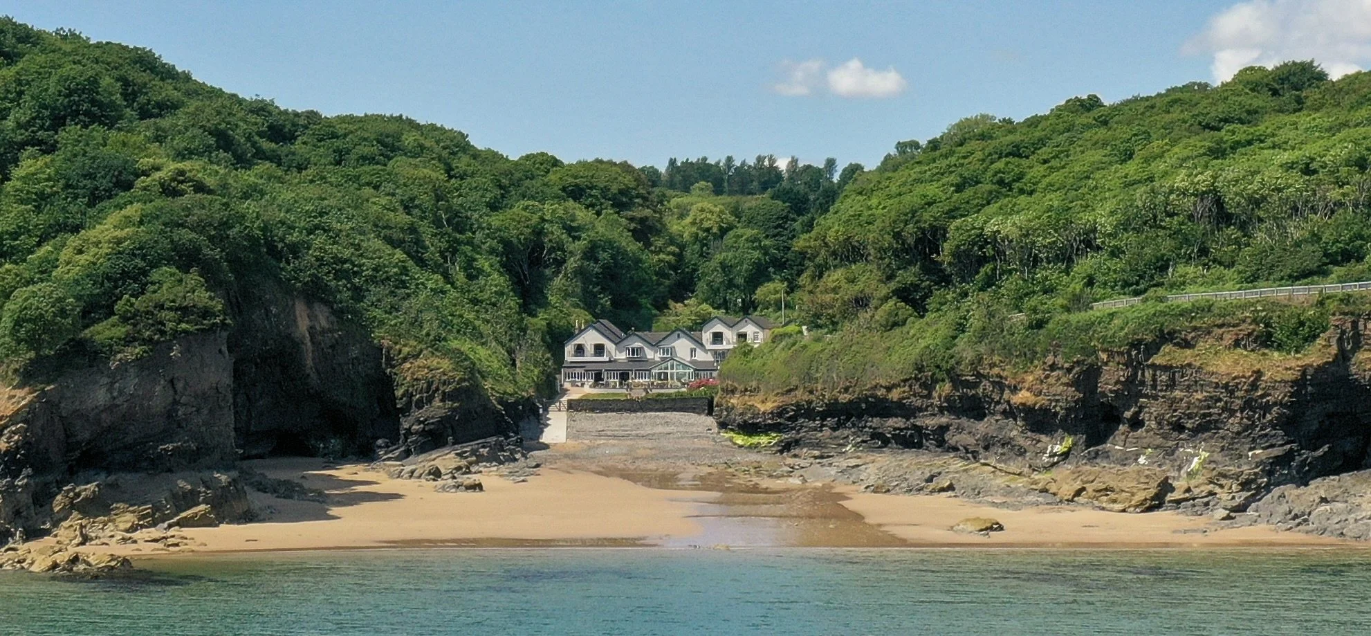 House situated on a hillside with green trees, rocky cliffs, sandy beach, and calm water in the foreground.