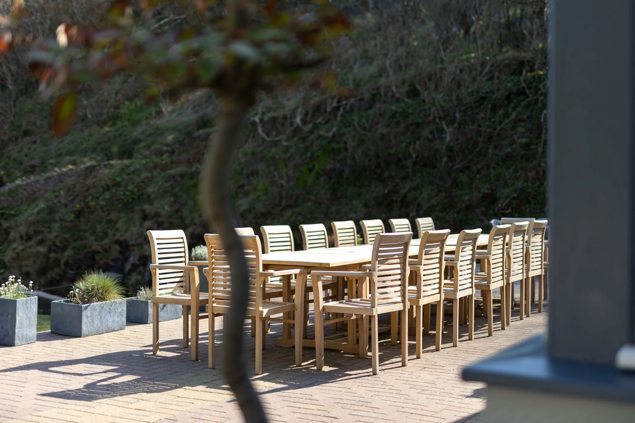Empty outdoor patio with a long wooden dining table and matching chairs, surrounded by potted plants, with a green hillside in the background.