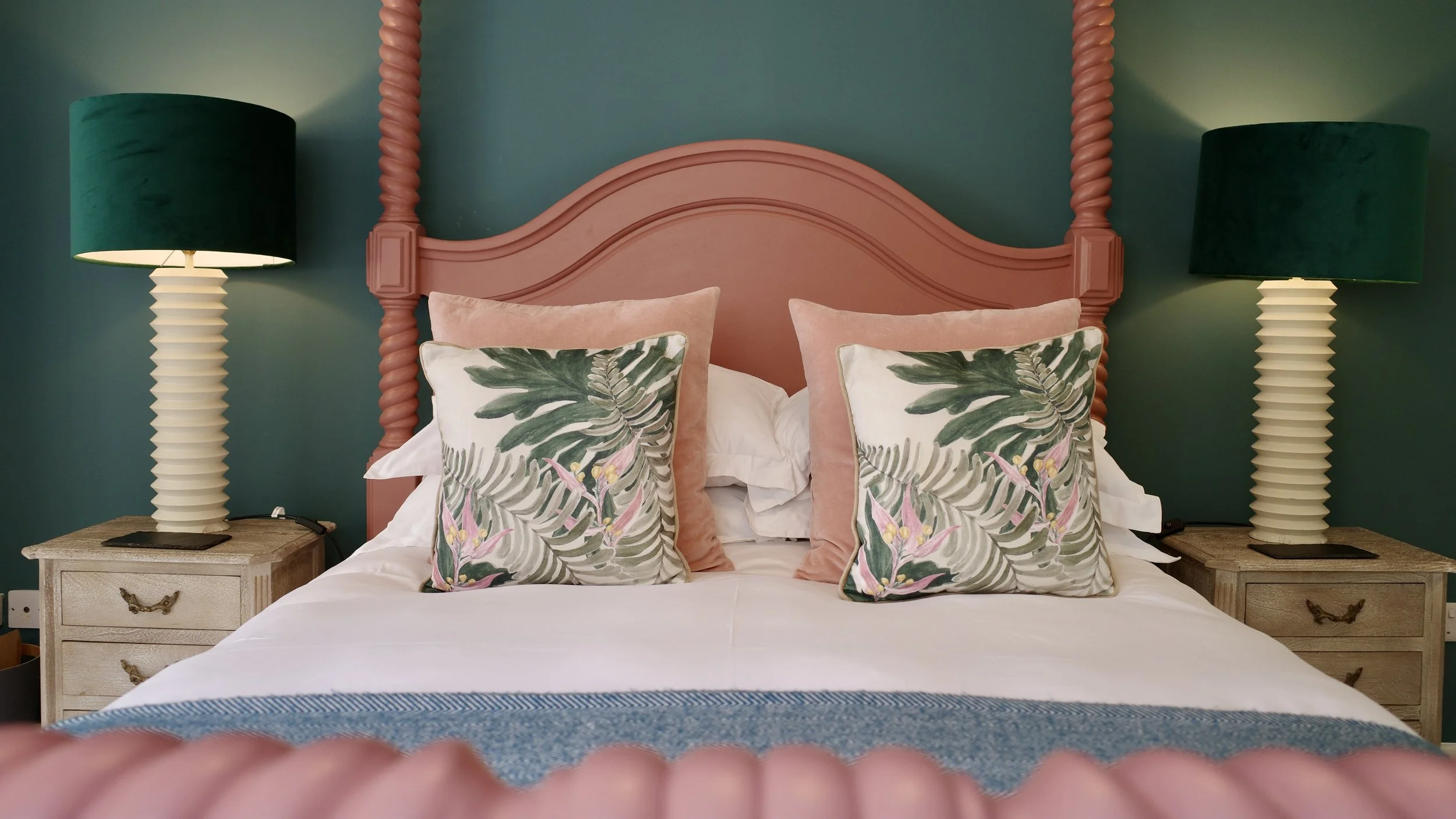 Bedroom with a pink headboard, two green lamps, and pillows with tropical leaf and floral designs.