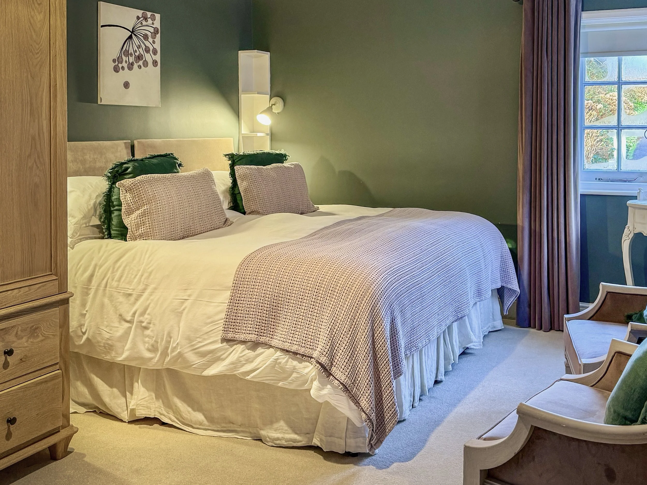 A cozy bedroom with a large bed made up with cream-colored bed linens, plaid pillows, and a blanket. There is a wooden nightstand, a modern white lamp, and a picture of a botanical on the green wall. A window with pink curtains reveals an outdoor scene with greenery. Two wooden chairs with green cushions are positioned near the window.