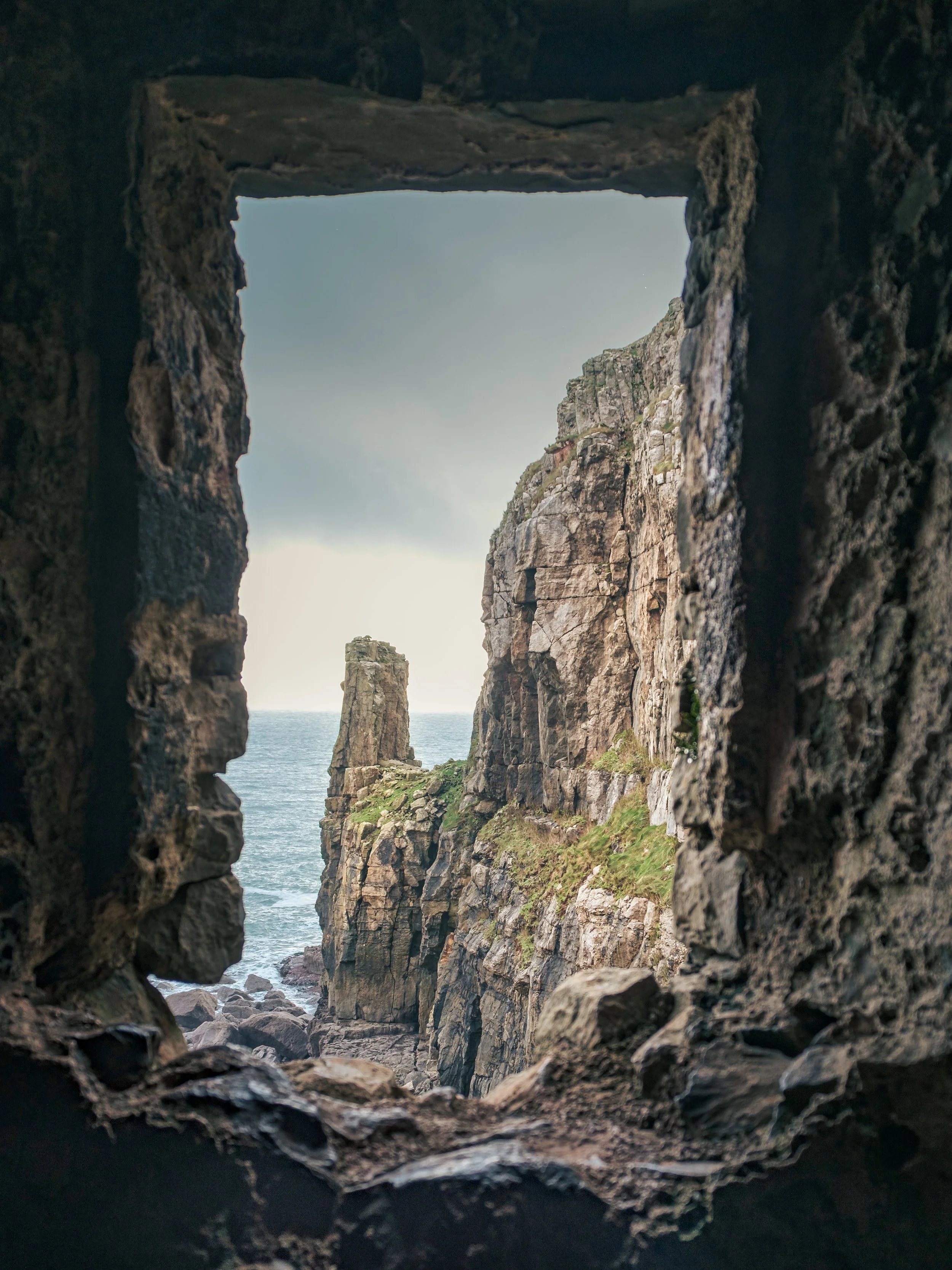 View through a rocky opening towards tall cliffs, the ocean, and a cloudy sky.