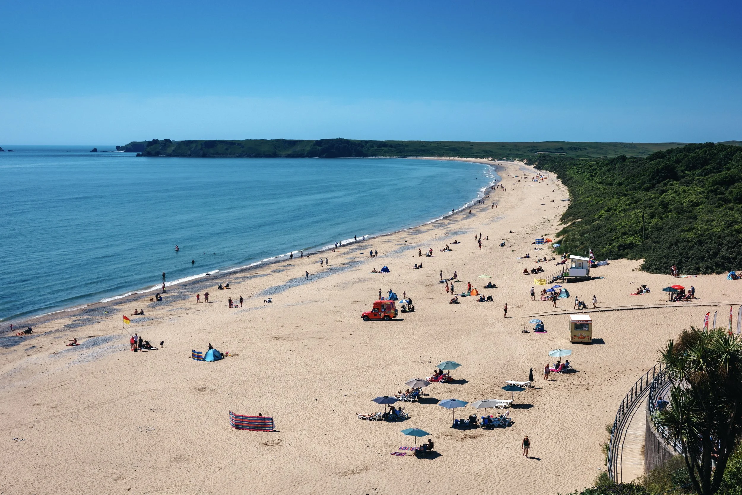 A wide view of a sandy beach with many people relaxing, swimming, and walking along the shoreline. There are umbrellas, tents, and a small ice cream stand on the beach. The ocean is calm with a few people in the water, and a green landmass is visible in the distance under a clear blue sky.