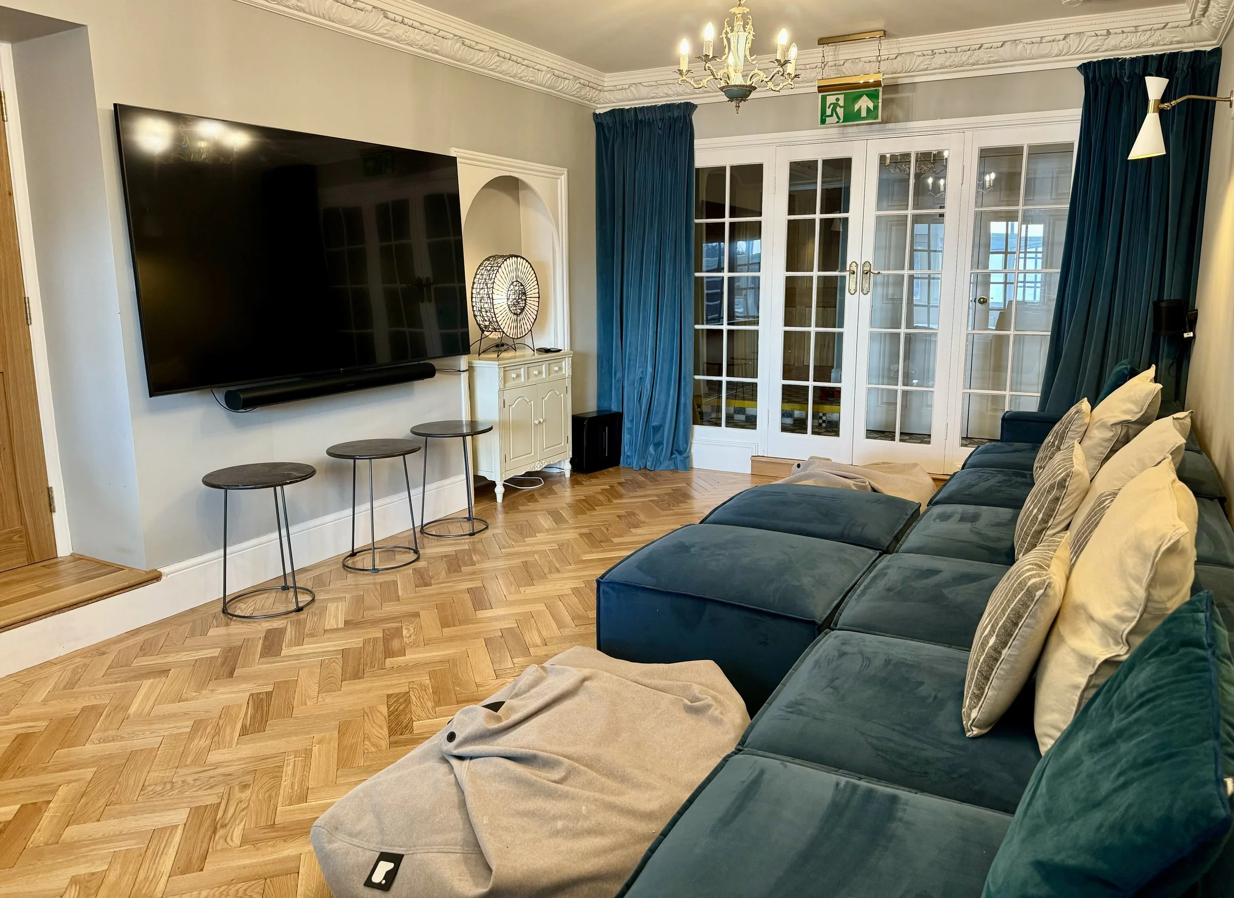 A living room with a flat-screen TV mounted on the wall, a small white cabinet below the TV, a lamp with a decorative wire shade on the cabinet, a blue velvet couch with throw pillows, wooden flooring, and large glass double doors with blue curtains leading to a balcony or outside area. There is an emergency exit sign above the doors and a chandelier hanging from the ceiling.