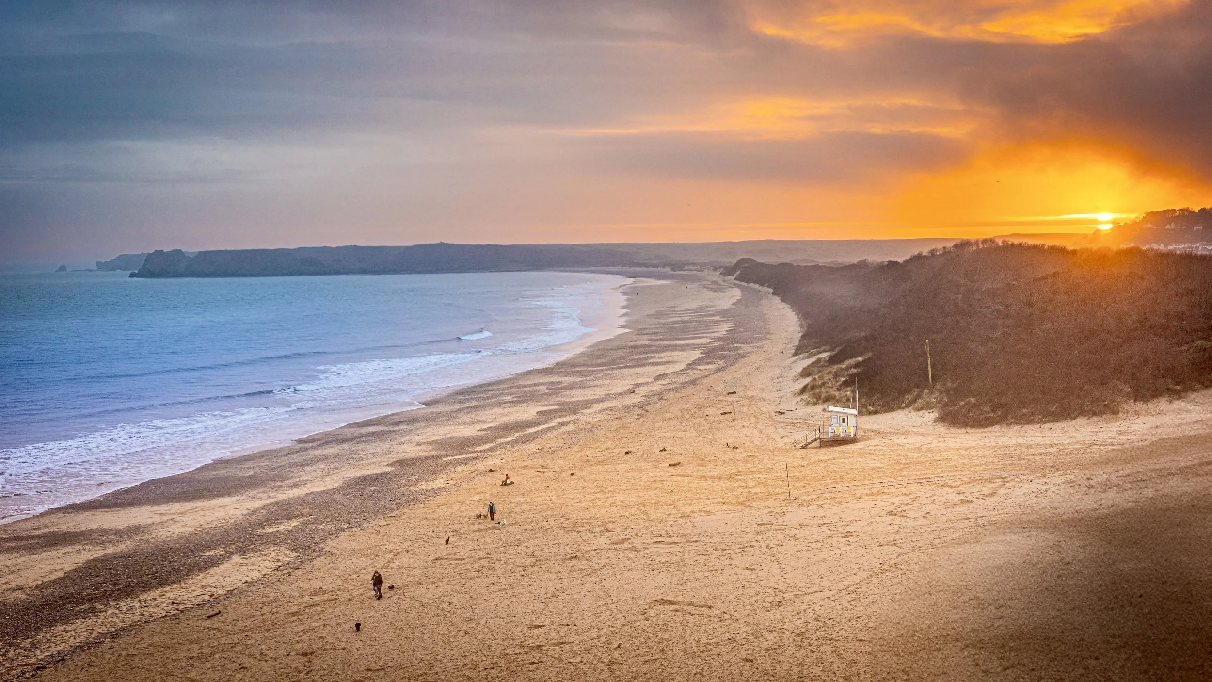 A beach at sunset with a few people walking along the sand and a small lifeguard station near the shoreline. The sky is partly cloudy with the sun setting on the horizon, casting a warm glow over the scene.
