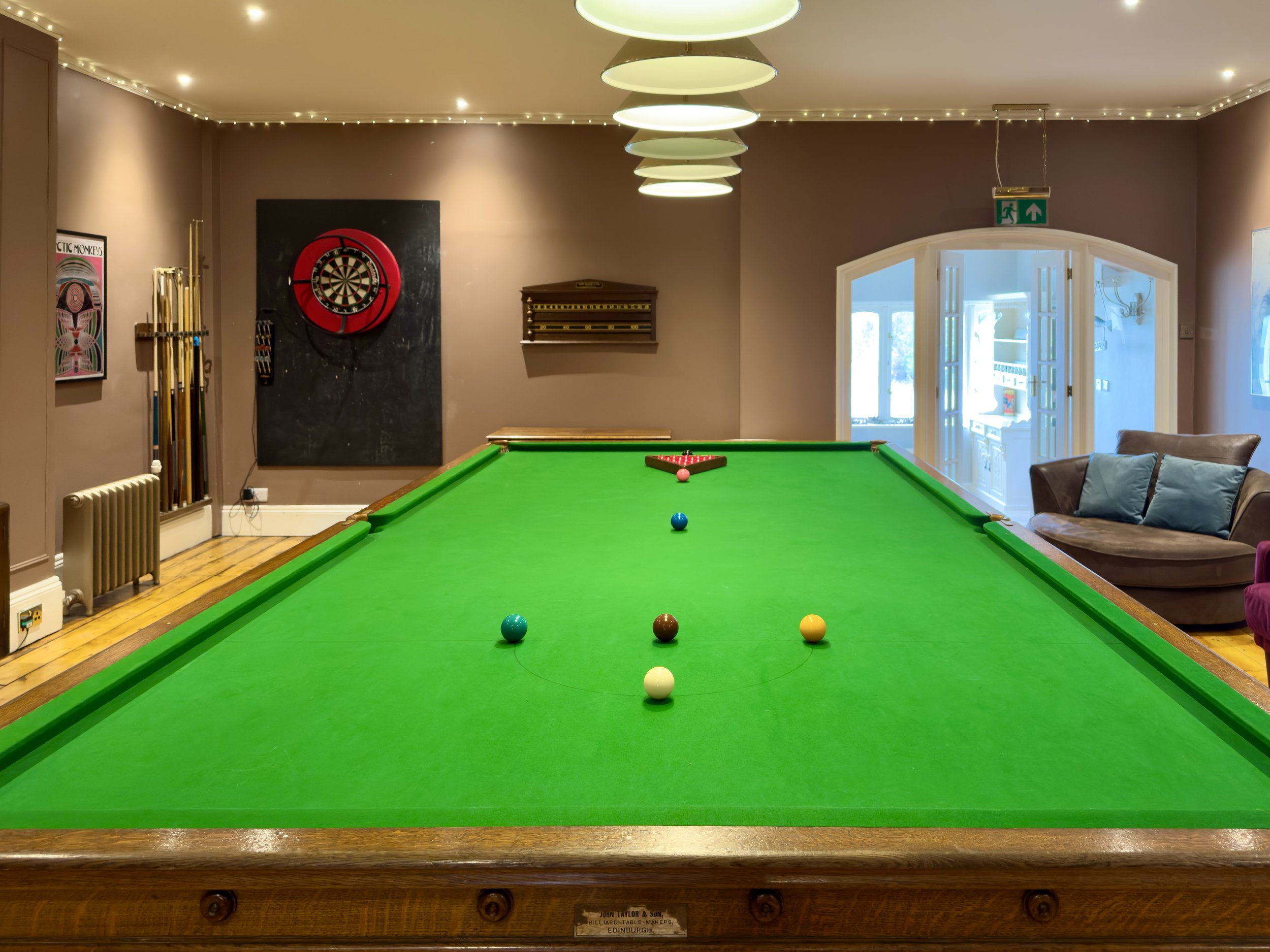 Indoor room with a green pool table, a dartboard on the wall, billiard cues on a rack, a dartboard cabinet, a sofa, and large windows with a glass door, illuminated by ceiling lights and decorated with string lights.